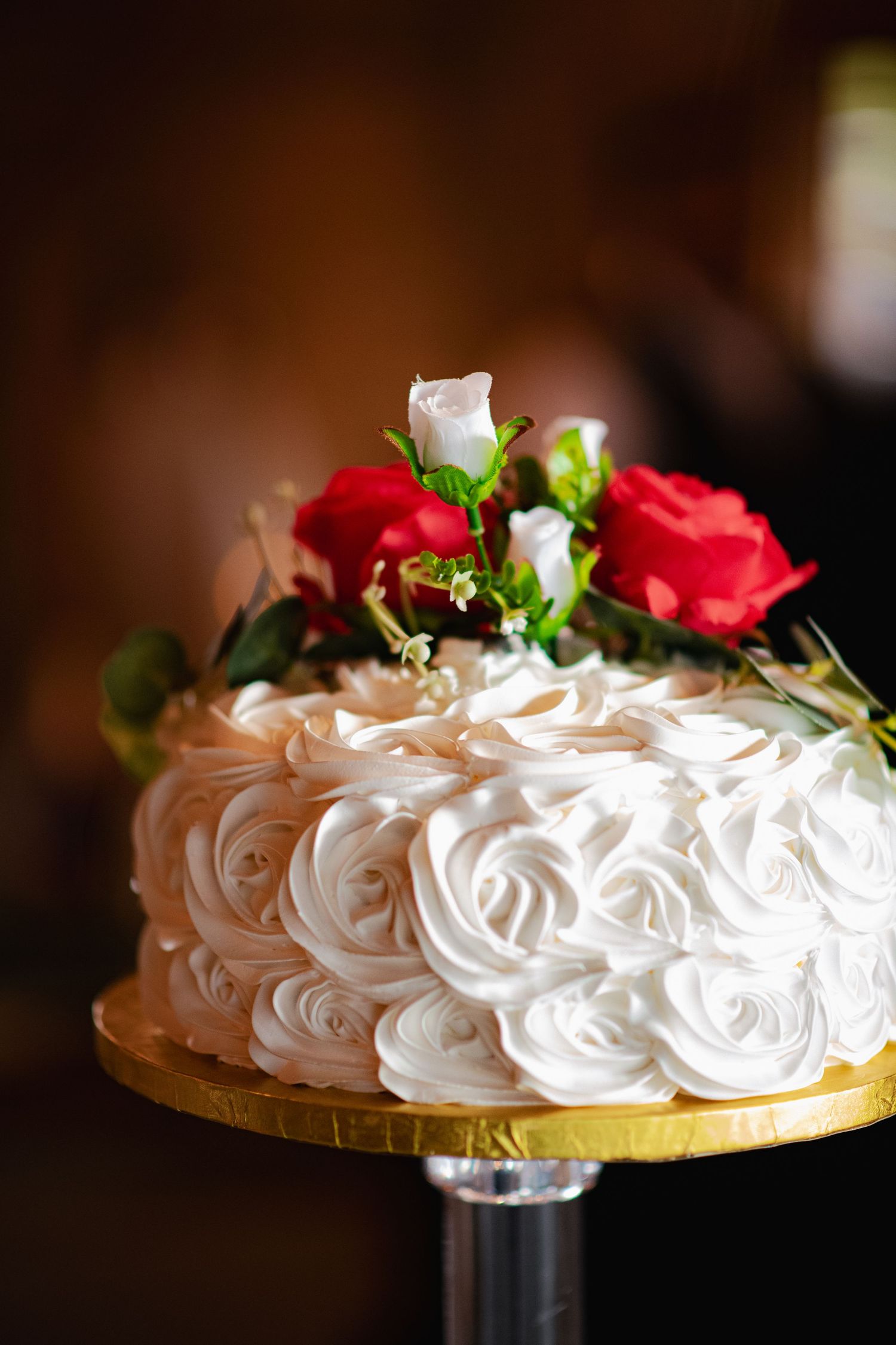 White frosted wedding cake decorated with rosettes and red roses on a gold cake stand.