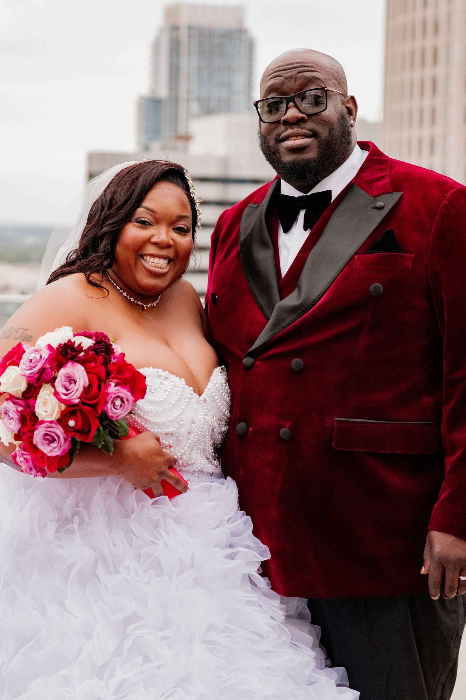 A couple poses against a city backdrop, with the bride in white and groom in burgundy velvet.