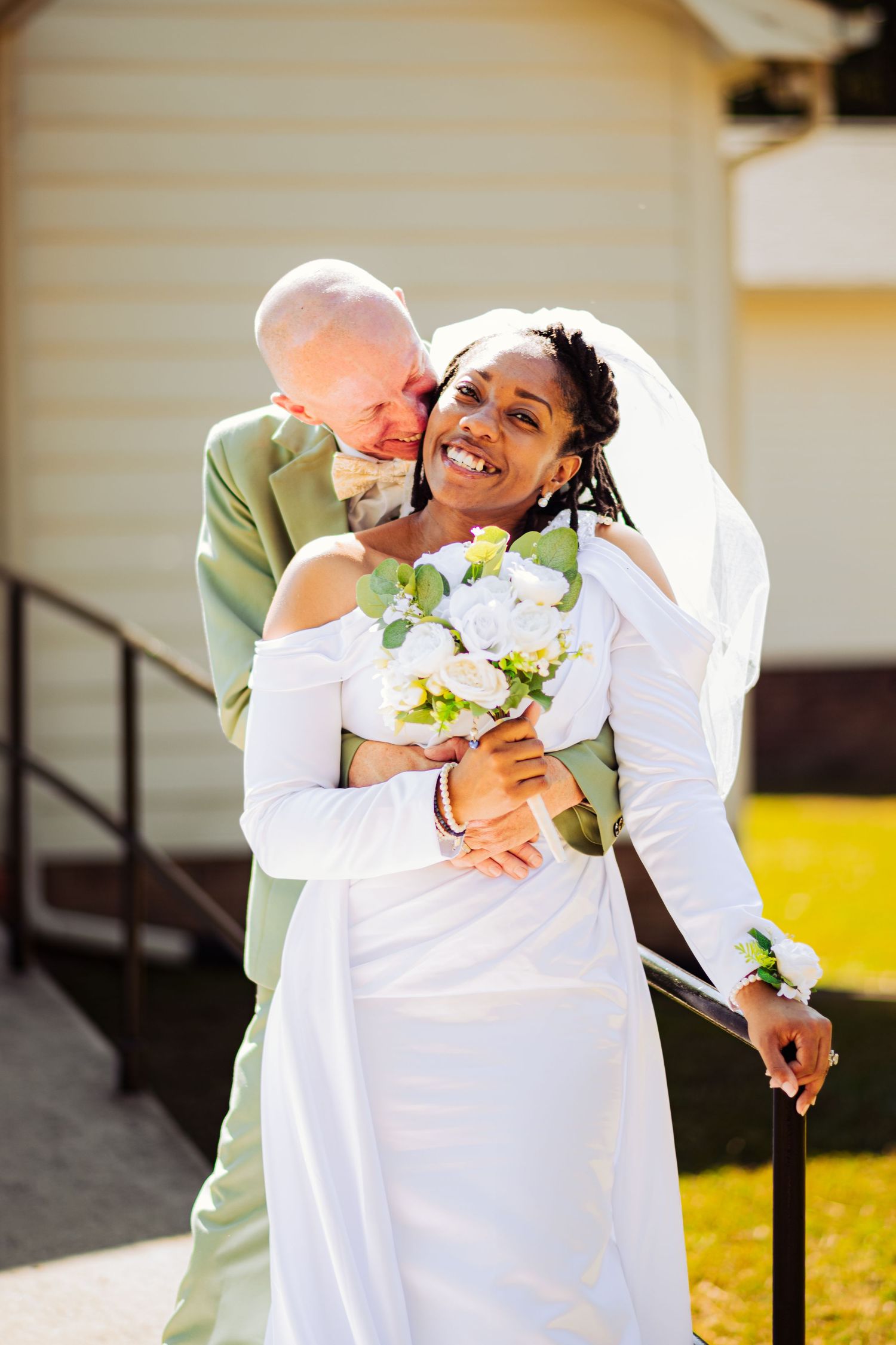 Joyful wedding couple embracing and laughing together outdoors in white attire.