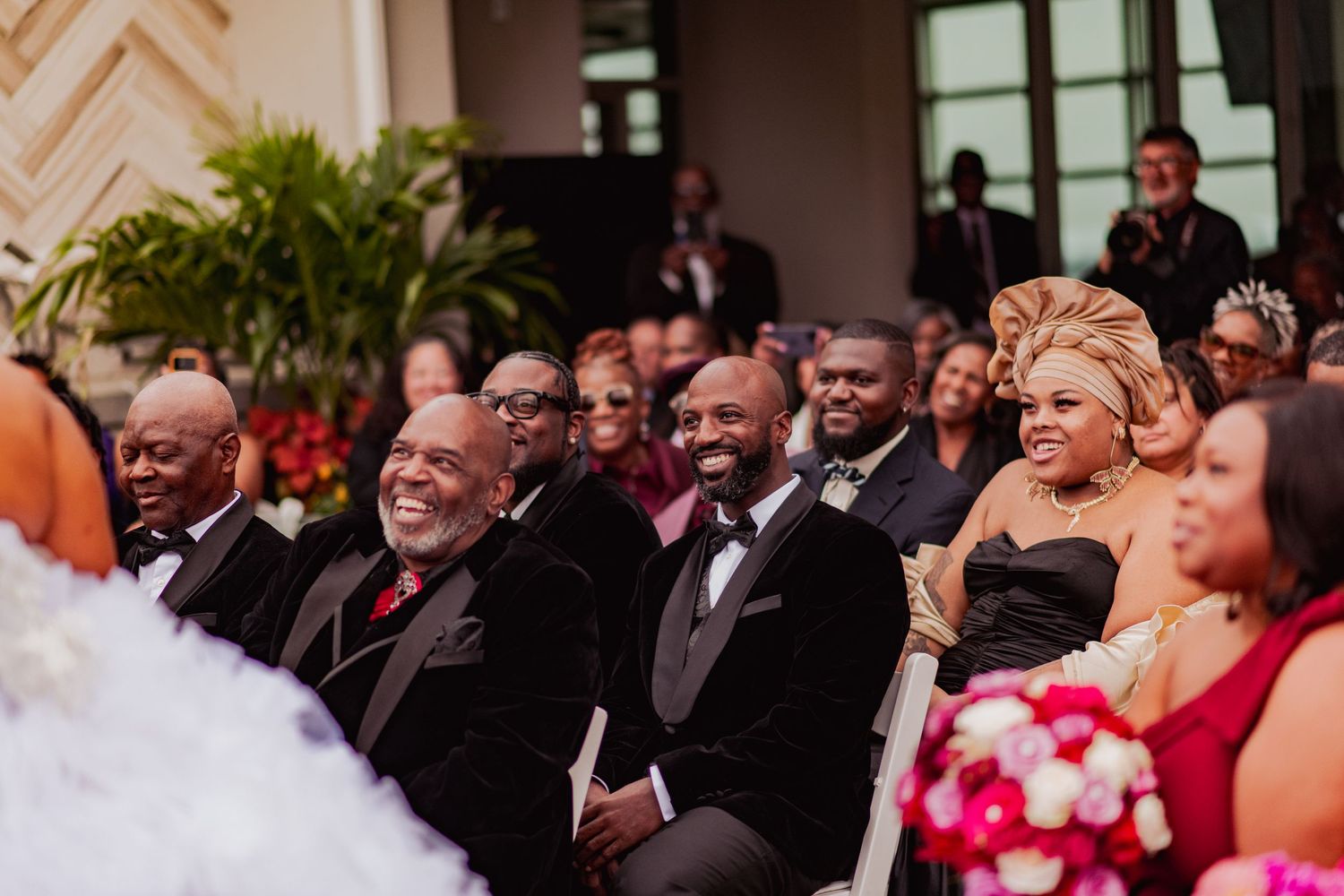 Wedding guests smile and react emotionally during a ceremony while seated in rows with decorative plants in the background.