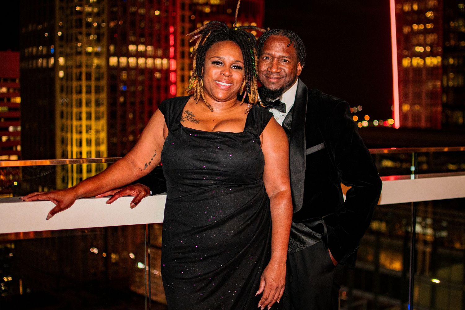 Elegant couple poses against nighttime city skyline at formal event.