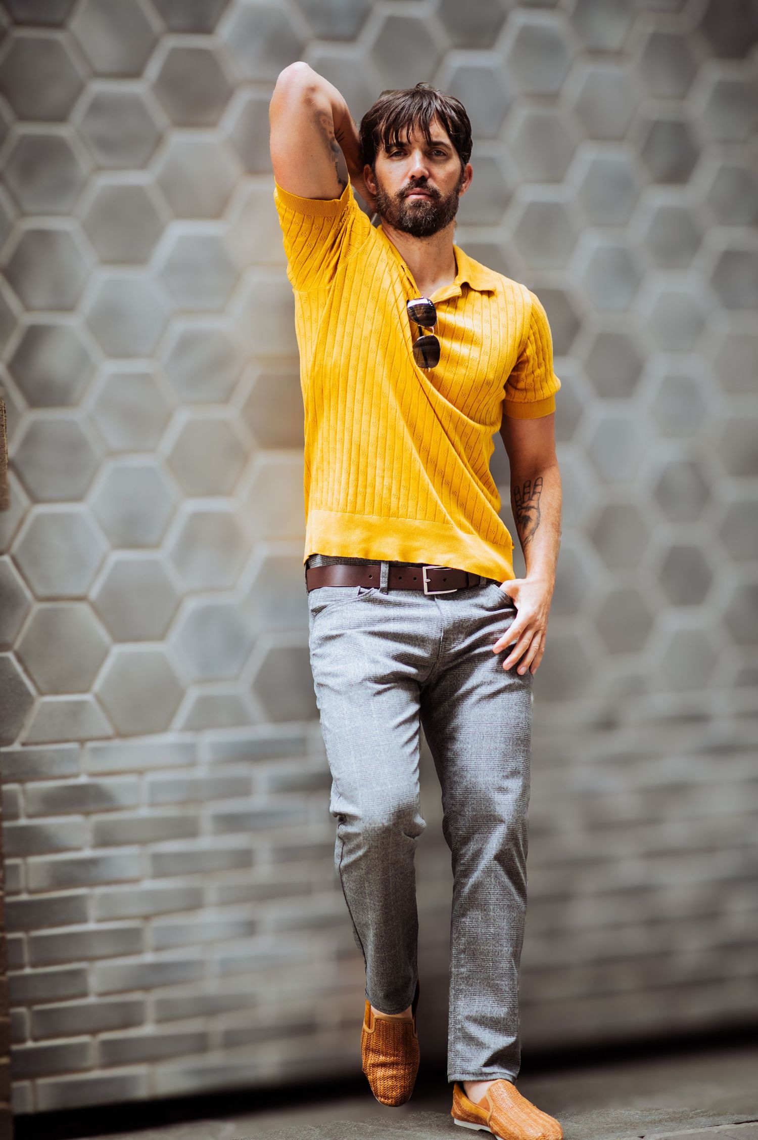 A person in a yellow polo shirt and gray pants poses against a geometric wall pattern.