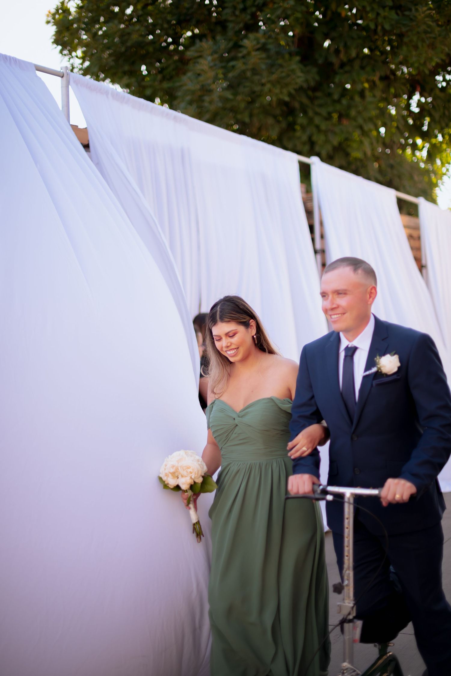 Wedding party member in sage green dress walks arm in arm with groomsman against white backdrop.