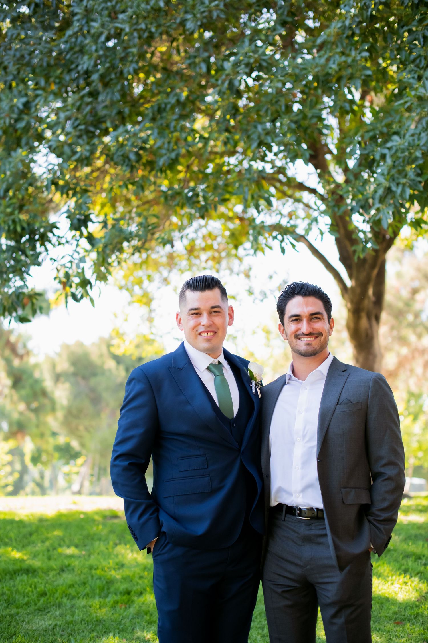 Two friends standing together outdoors under green trees on a sunny day.