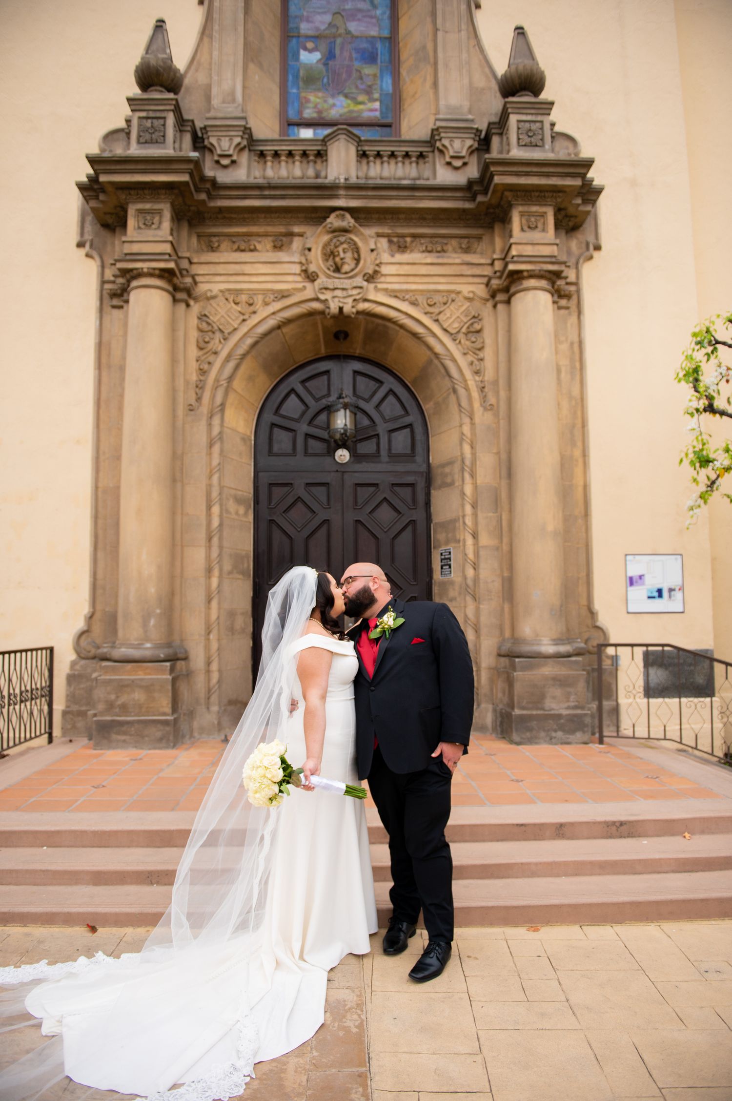 A wedding couple poses in front of a grand church entrance with ornate stone architecture and wooden doors.