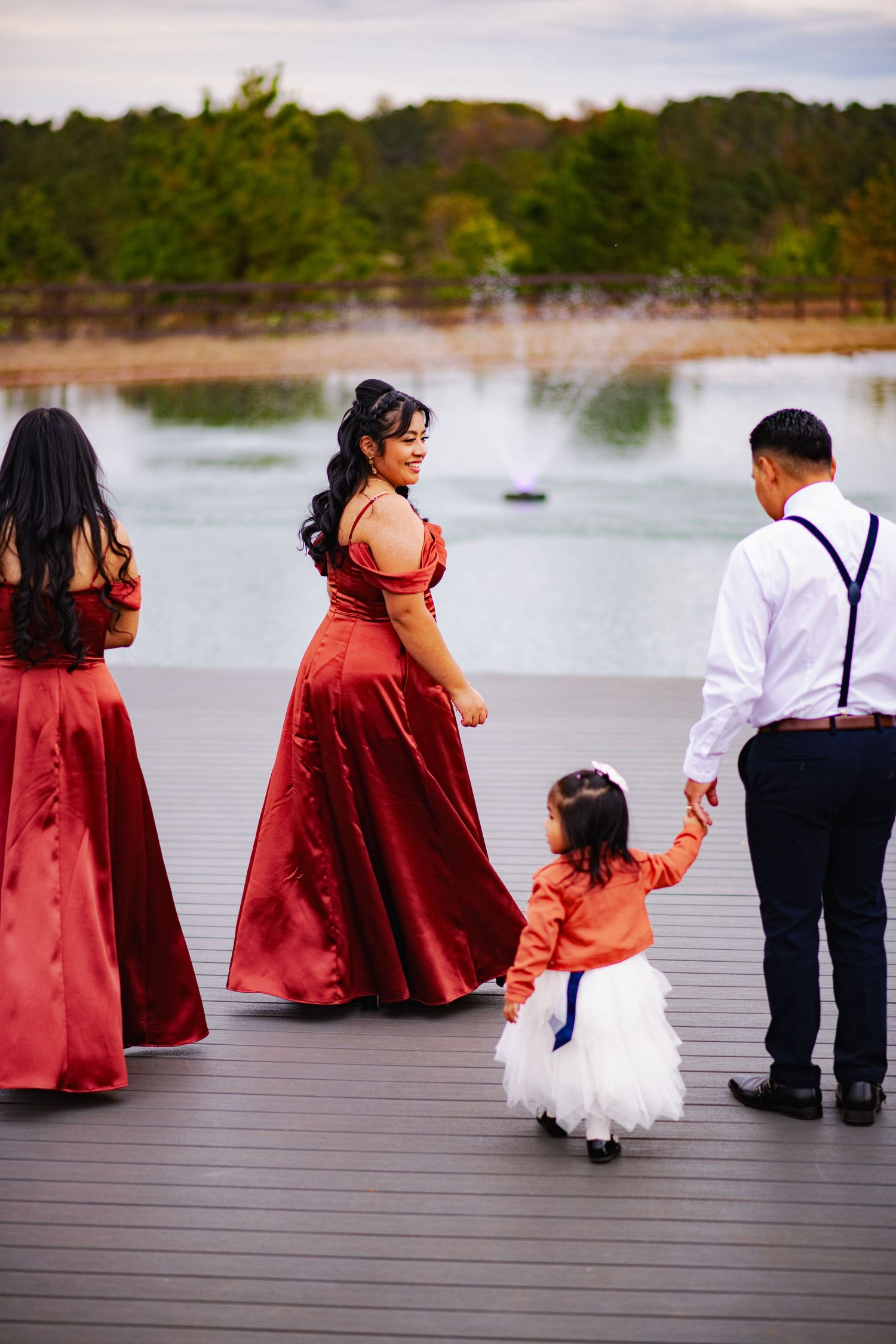 Wedding party in red and formal attire poses on wooden dock by lake.