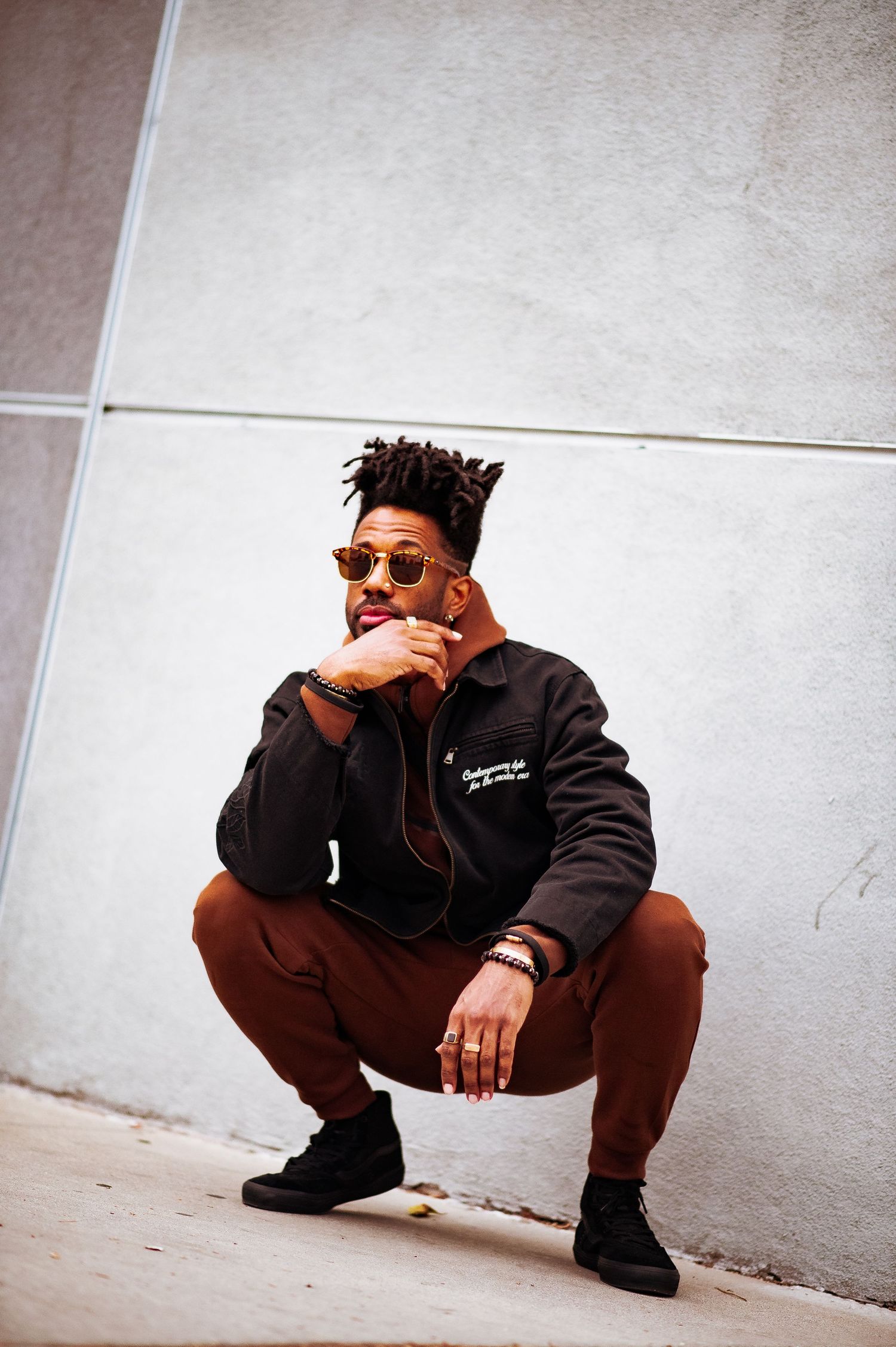 Figure in dark clothing and sunglasses crouches against white wall in stylish urban photoshoot.