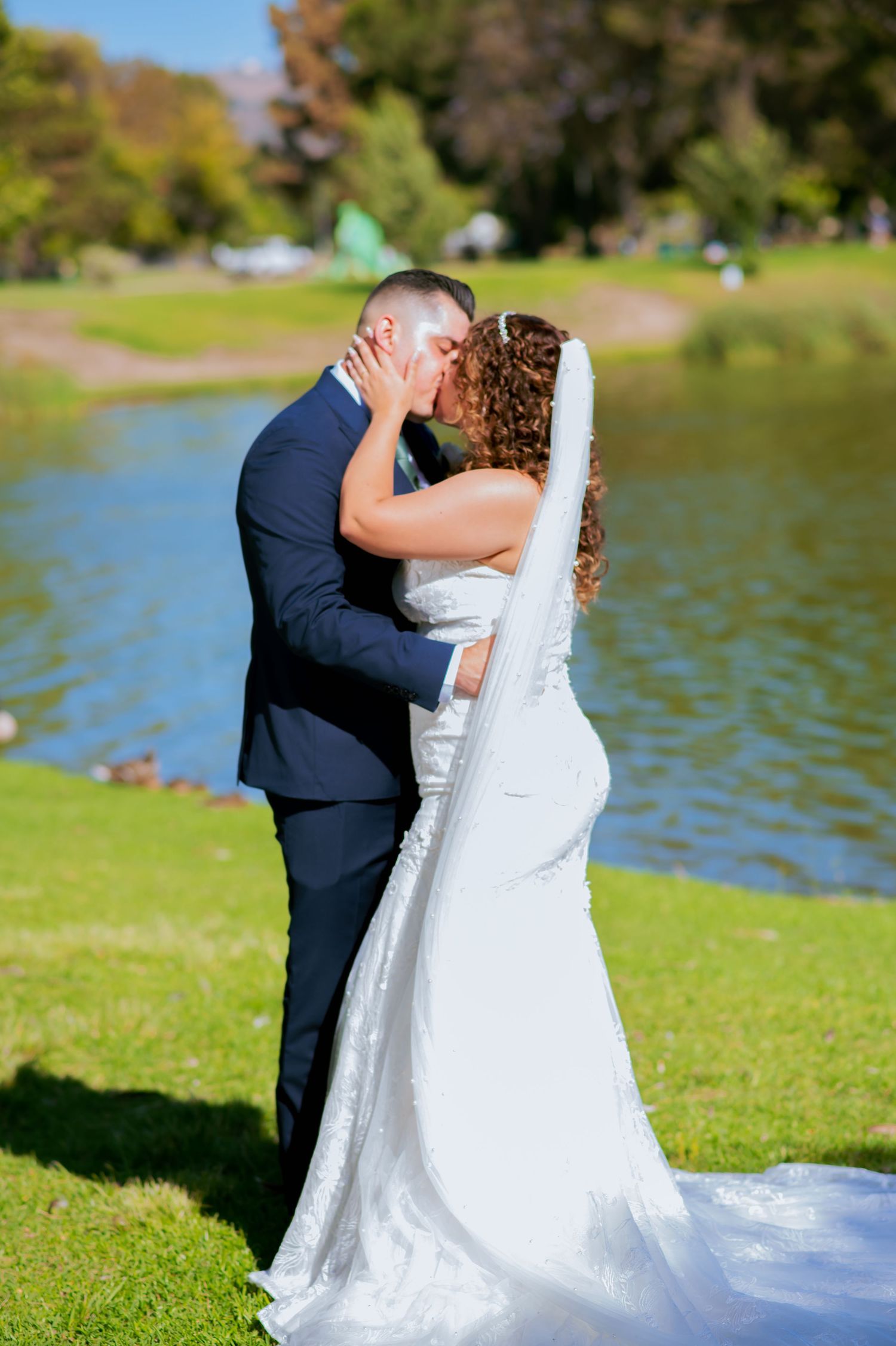 Newlyweds share intimate moment by lakeside with bride's white dress trailing on green grass.