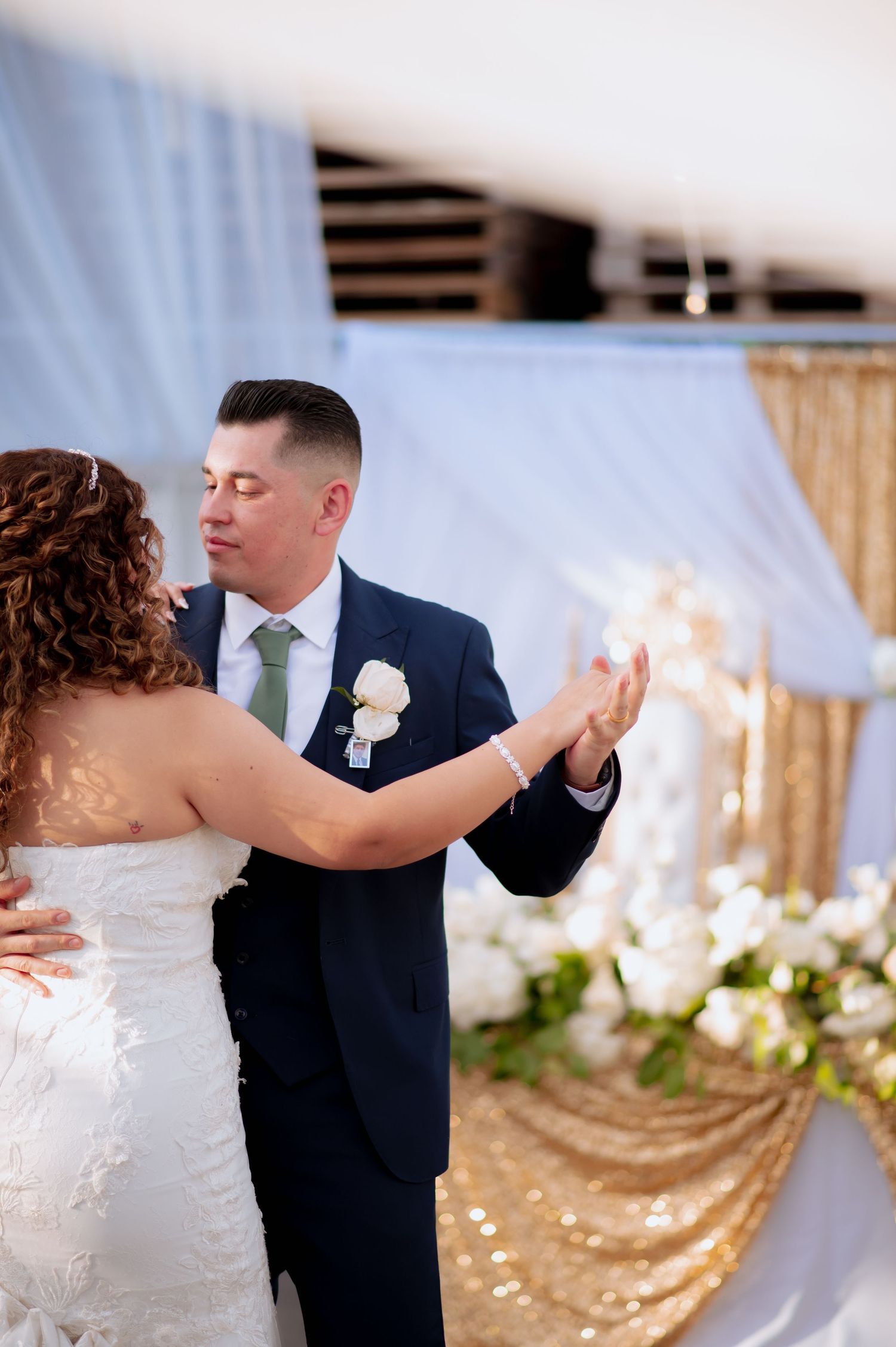 Bride in white gown and groom in navy suit dance together under white drapes and gold wedding decor.