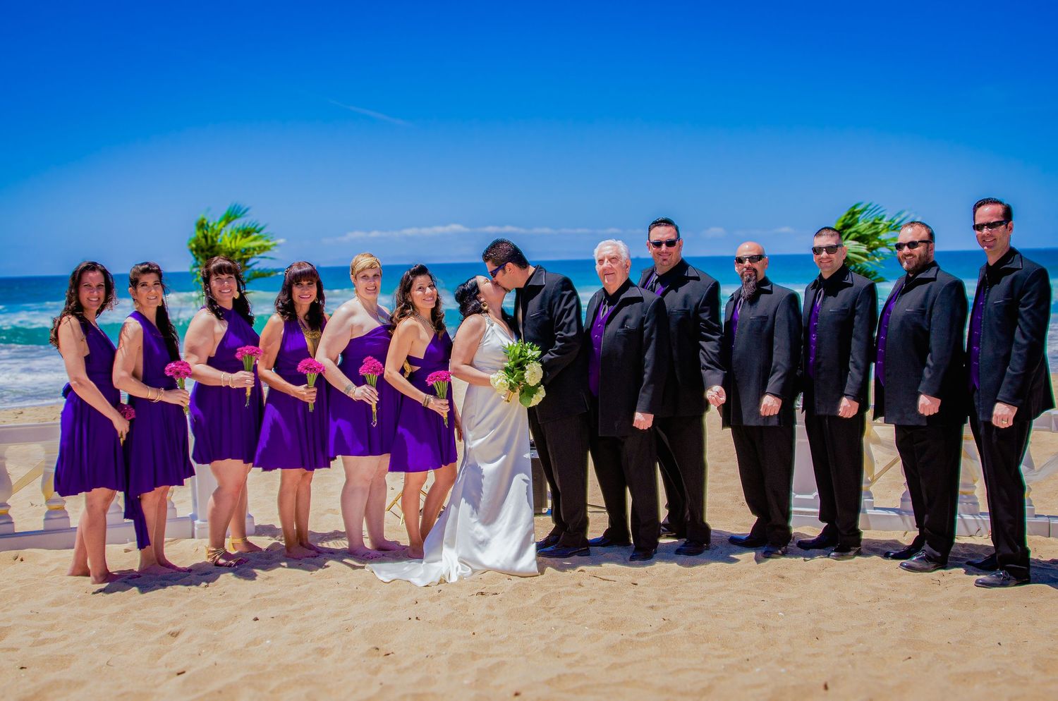 Wedding party lined up on beach wearing purple dresses and dark suits.
