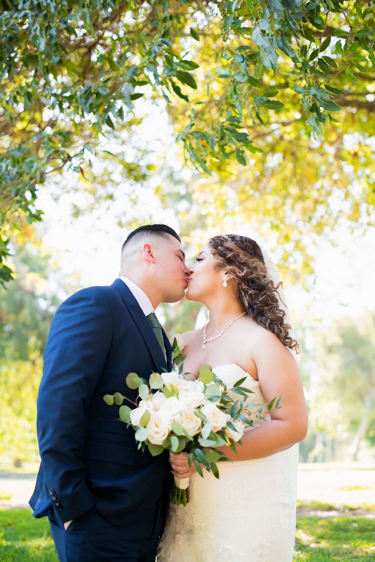 An intimate wedding portrait beneath sunlit trees with white roses and flowing veil.