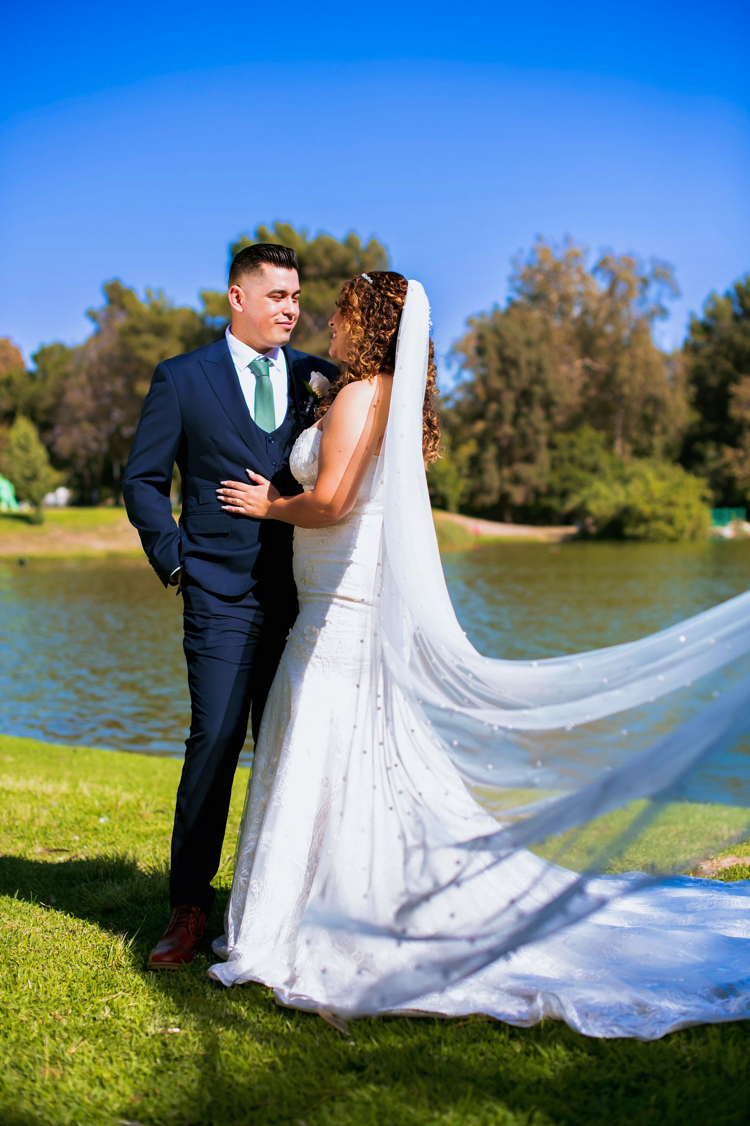 A romantic wedding couple stands by a lake while the bride's long white veil flows dramatically in the wind.