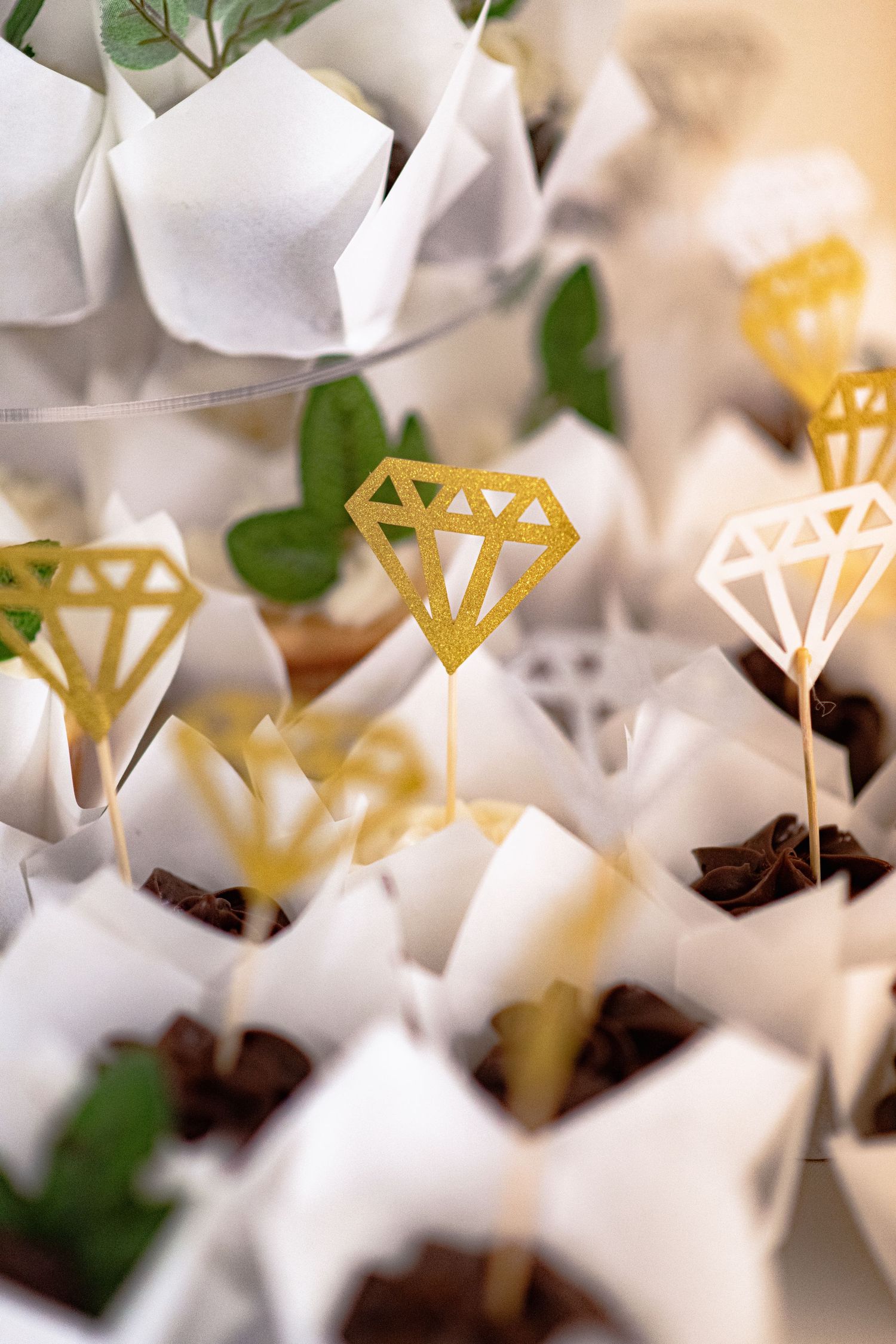 Gold diamond-shaped cake toppers decorate small desserts in white paper wrappers with greenery accents.