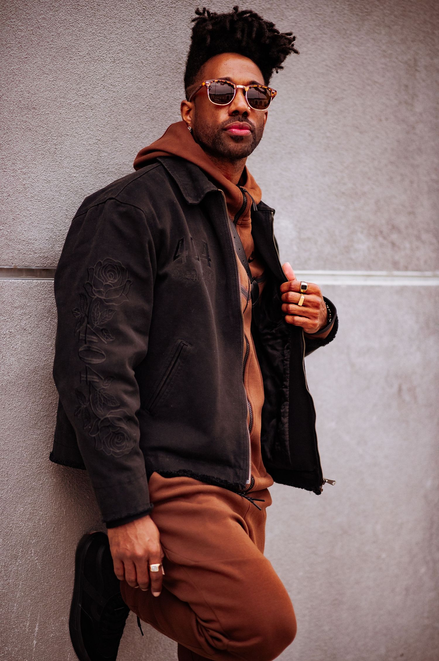 A person in sunglasses and black jacket poses against a wall wearing brown pants in a fashion-forward style.