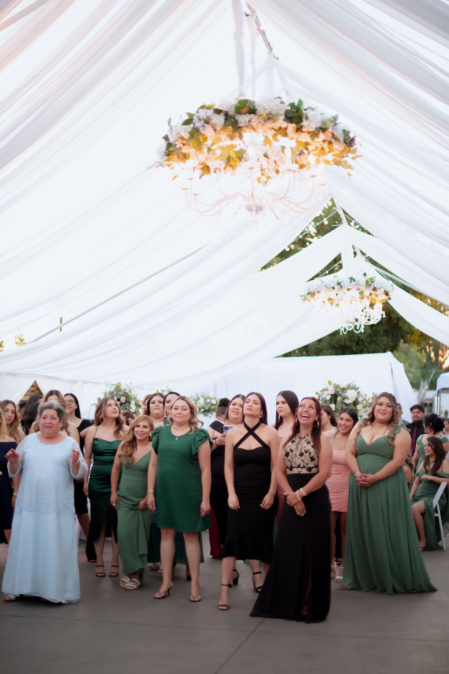 Wedding party members in green and black dresses dancing together under an elegant white tent.