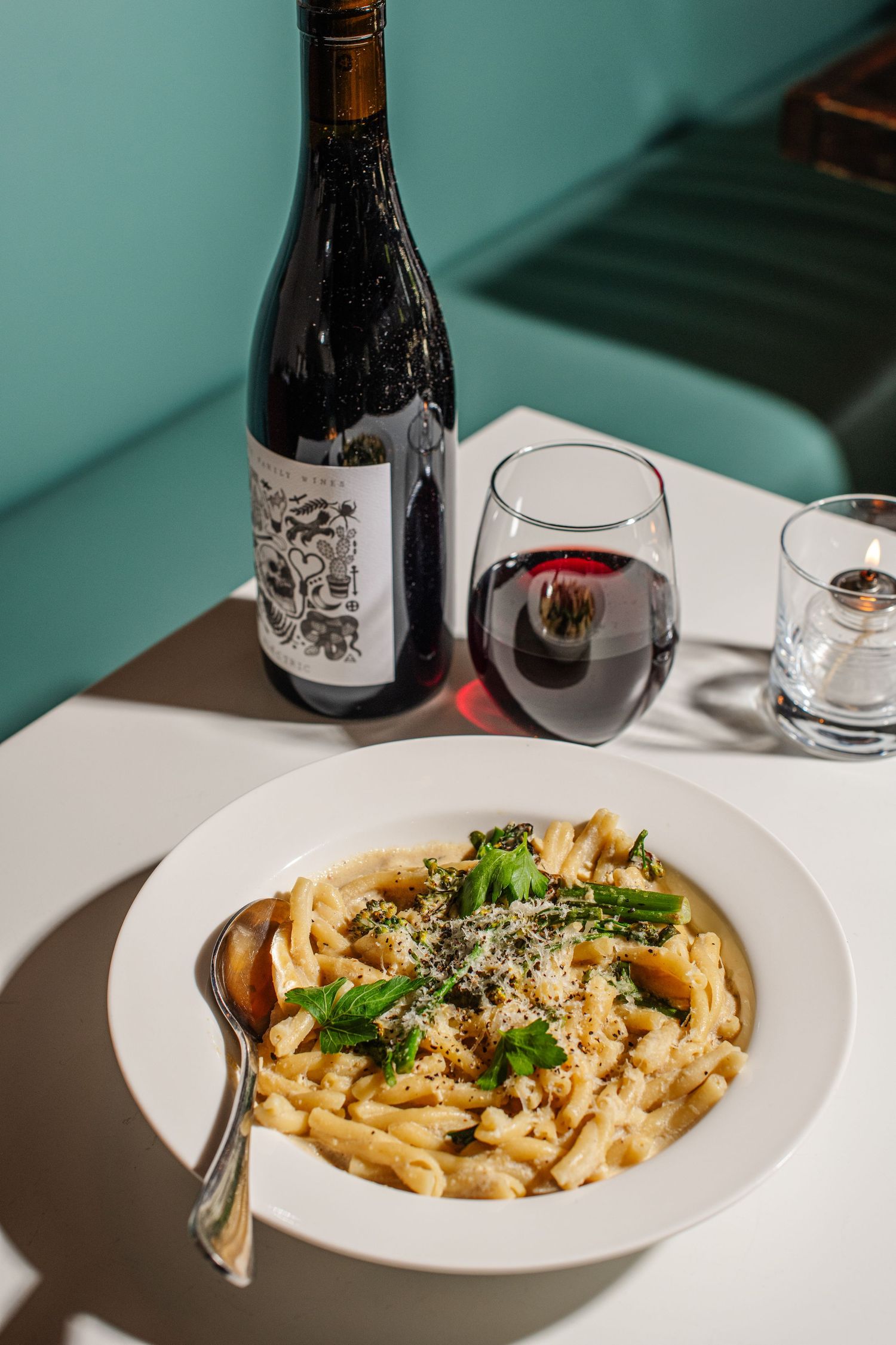 White bowl of creamy pasta with grated cheese and fresh herbs served alongside wine bottles on a turquoise background.