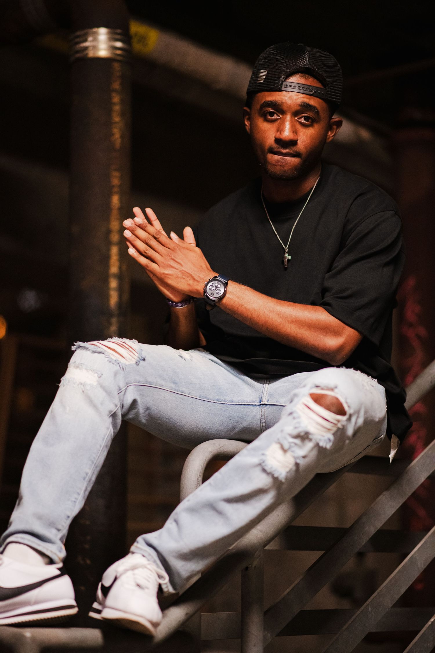 Christopher Riviere Stylish portrait session showing person in black attire and ripped jeans on industrial metal stairs in dark setting.