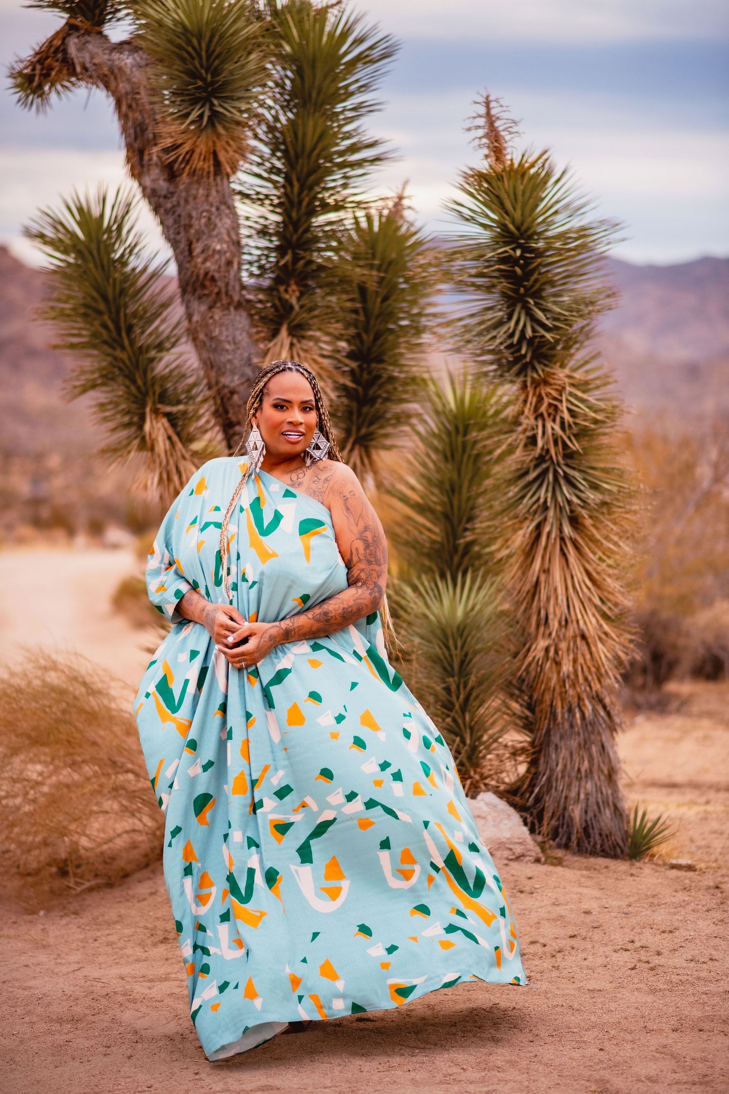 A flowing aqua dress with geometric patterns moves in the wind against desert mountains and yucca trees.