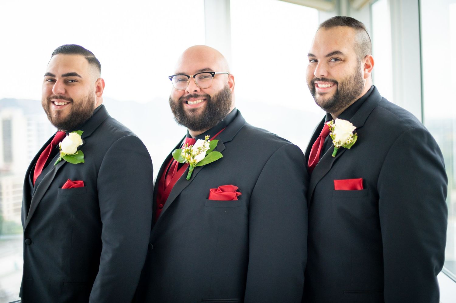 Groomsmen in black suits with red pocket squares and white boutonnieres pose together in a bright indoor setting.