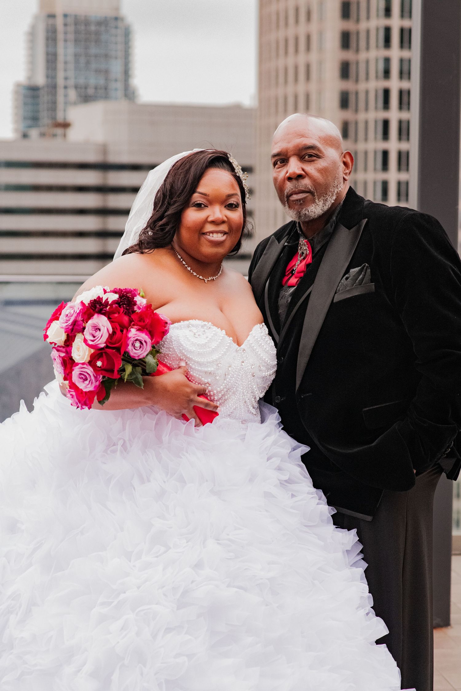 A bride in a white ballgown holds a pink bouquet while posing against an urban cityscape.