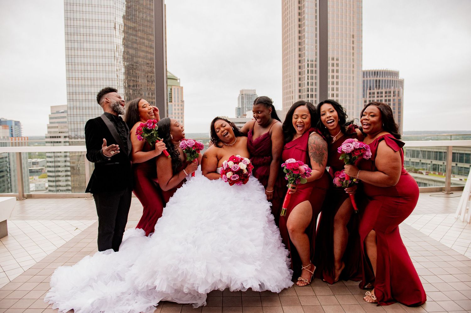 Large wedding party in red and black attire posing together on city rooftop terrace.