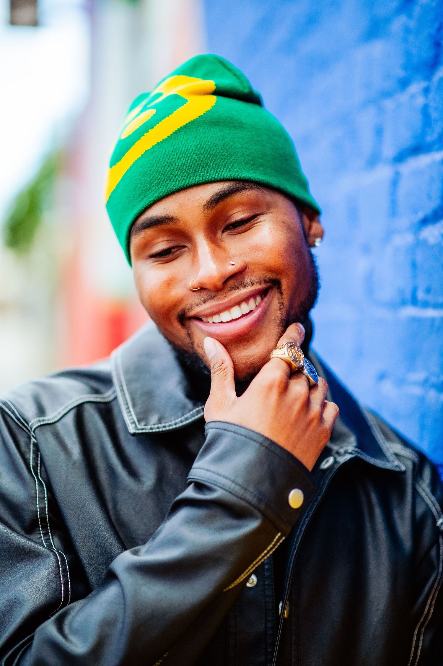 Person wearing a green beanie and black jacket laughs joyfully against a bright blue wall.