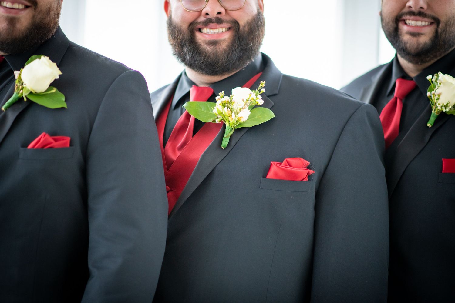 A group of groomsmen in black suits with red ties and pocket squares smile while wearing white boutonnieres.