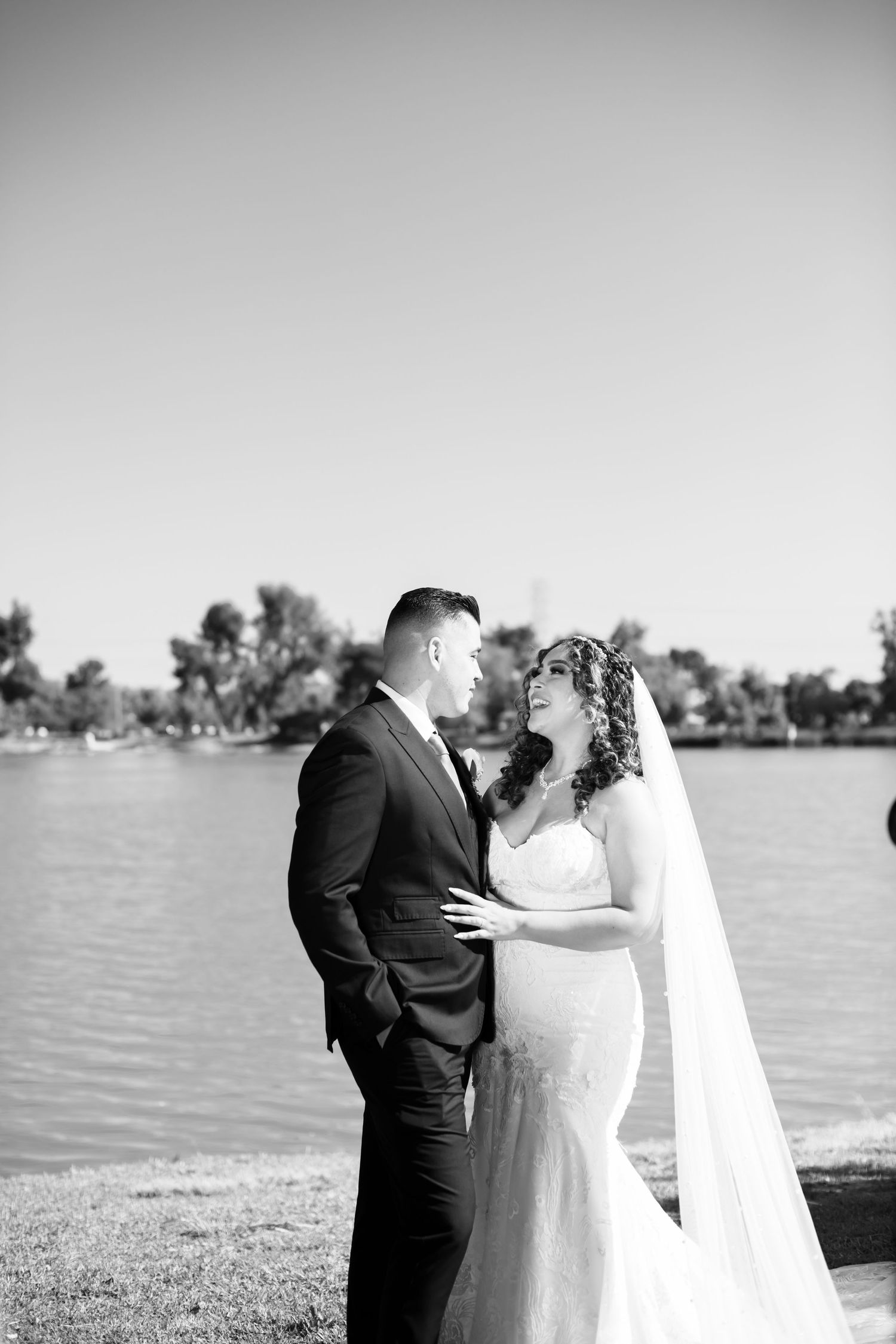 Black and white portrait of newlyweds embracing lakeside with trees in the background.