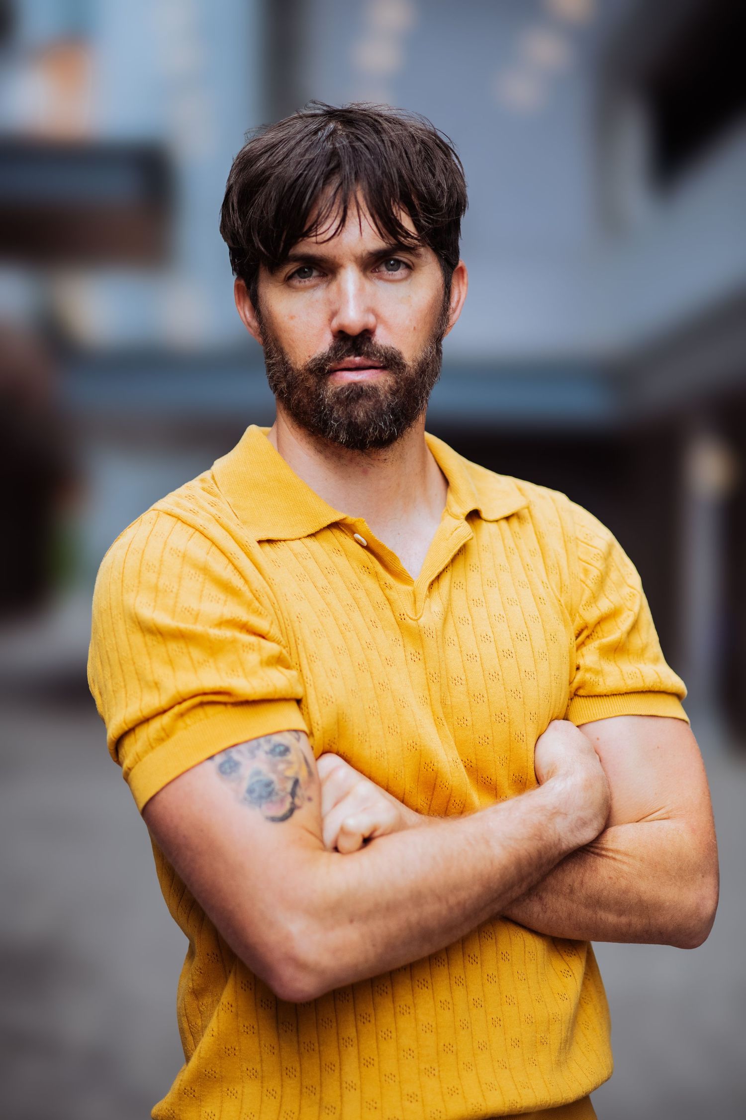 Confident pose with crossed arms wearing vibrant yellow shirt against blurred city background.