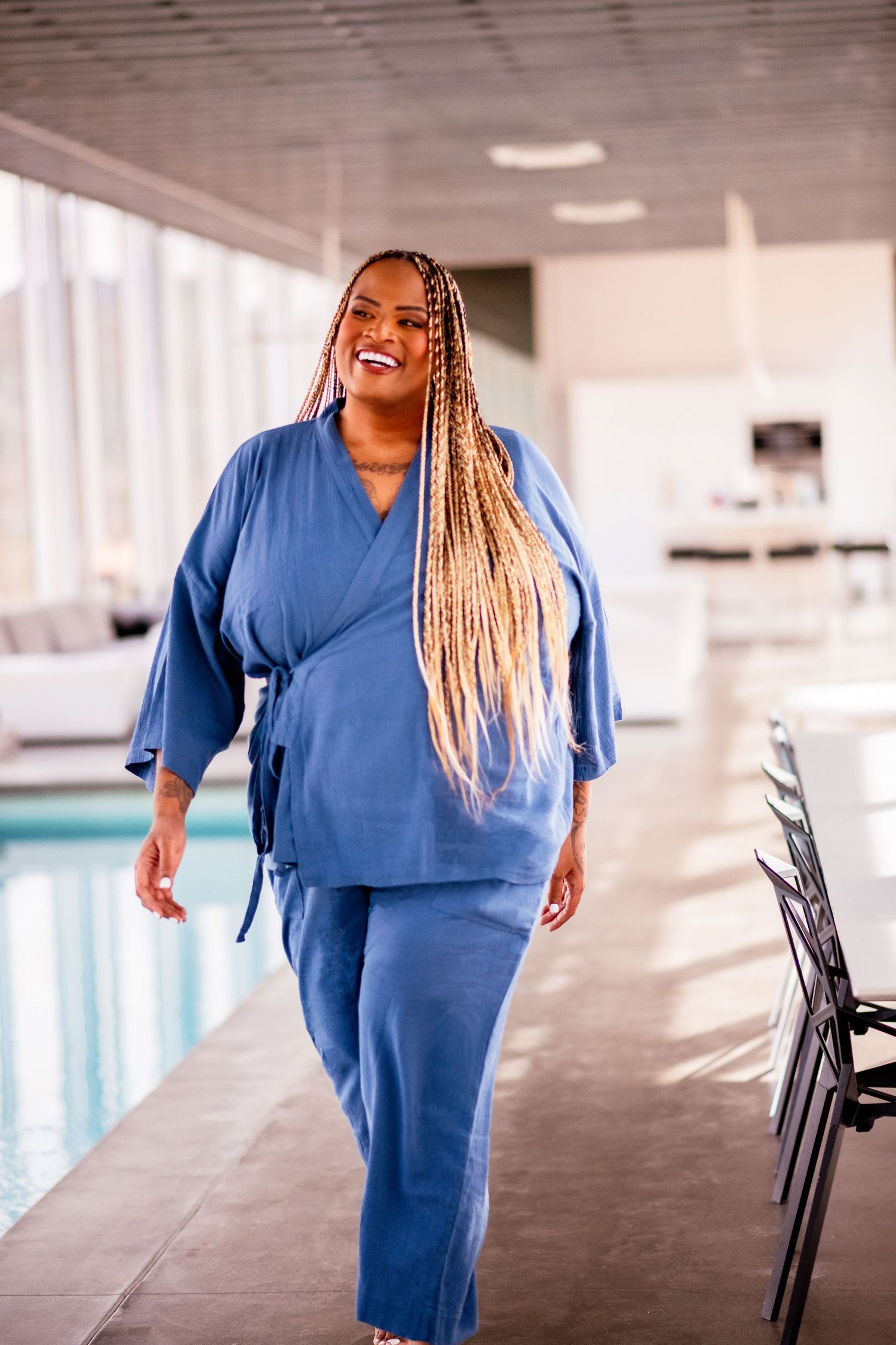 Confident person wearing blue linen spa robe and pants ensemble stands smiling in indoor poolside setting.
