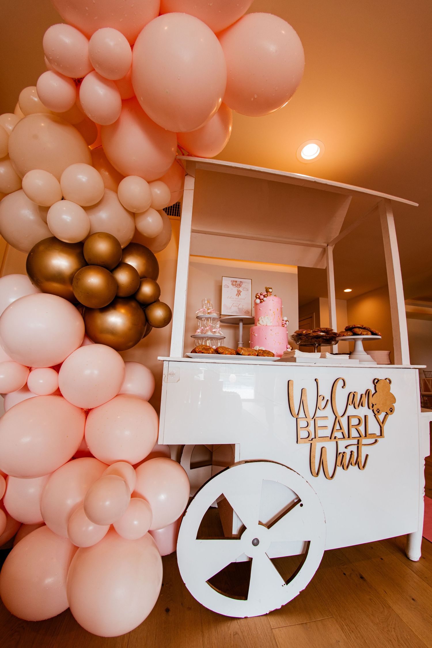 Elegant baby cart display with pink and gold balloon arch featuring desserts and treats for a celebration.