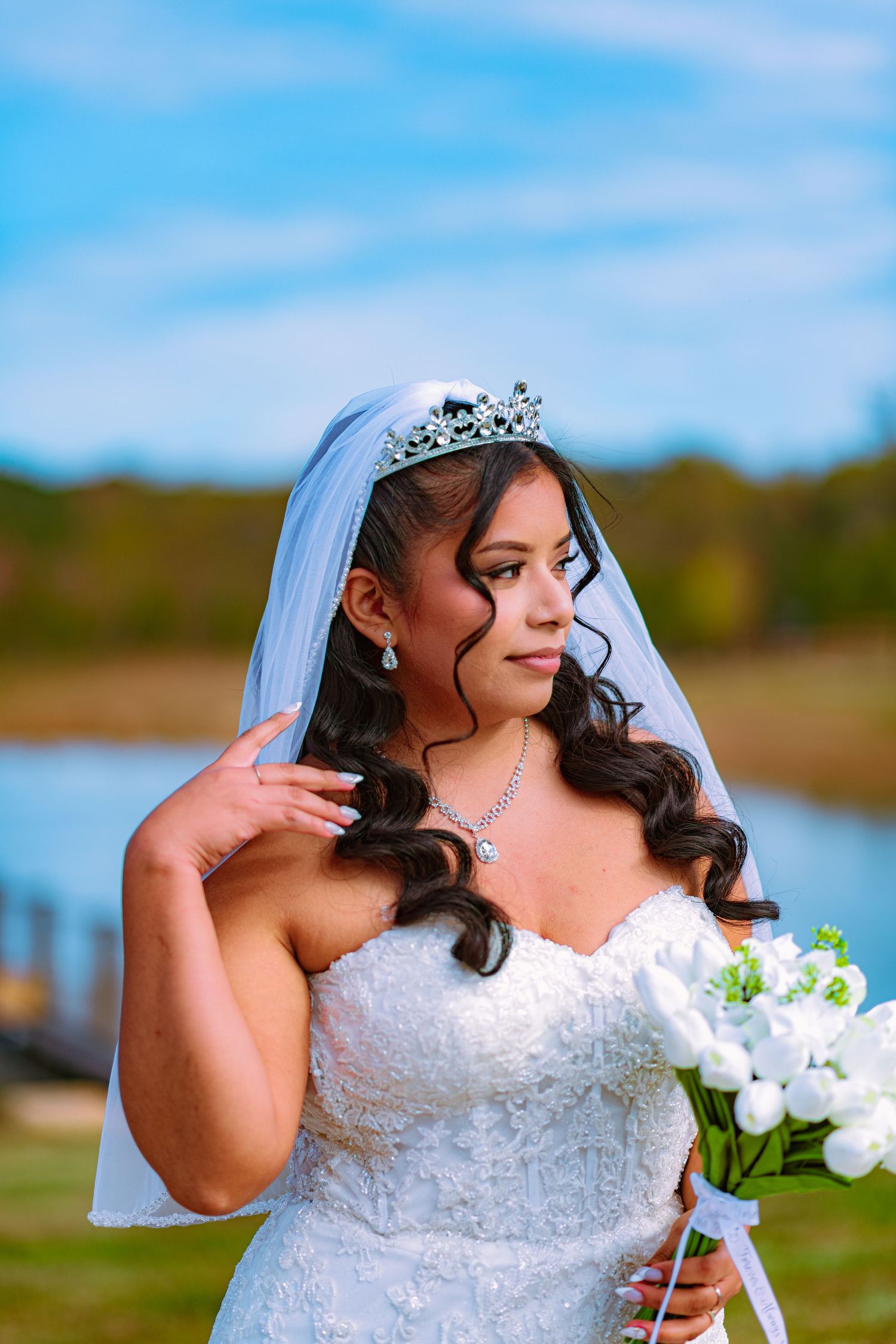 A bride in a white strapless gown with tiara and veil holds a bouquet against a blue sky backdrop.