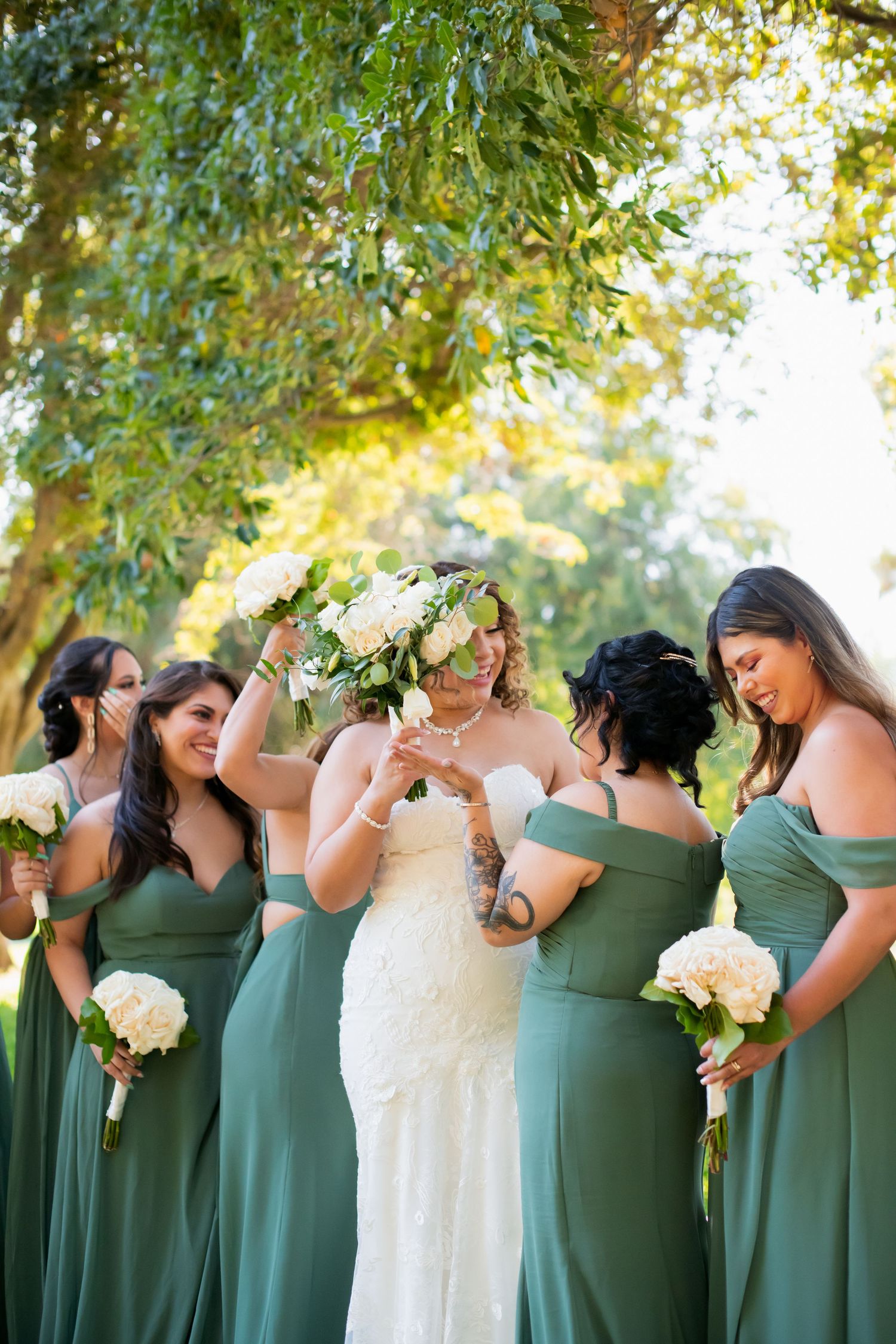 Wedding party in green bridesmaid dresses and white gown pose playfully in an outdoor setting.