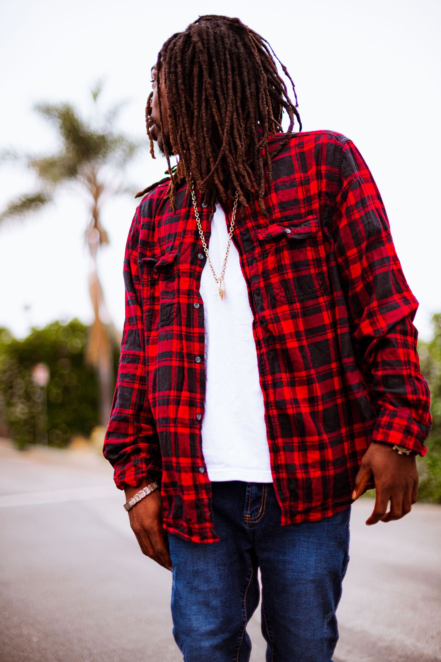 Person wearing a red and black buffalo plaid flannel shirt and blue jeans walks on a palm tree-lined street.