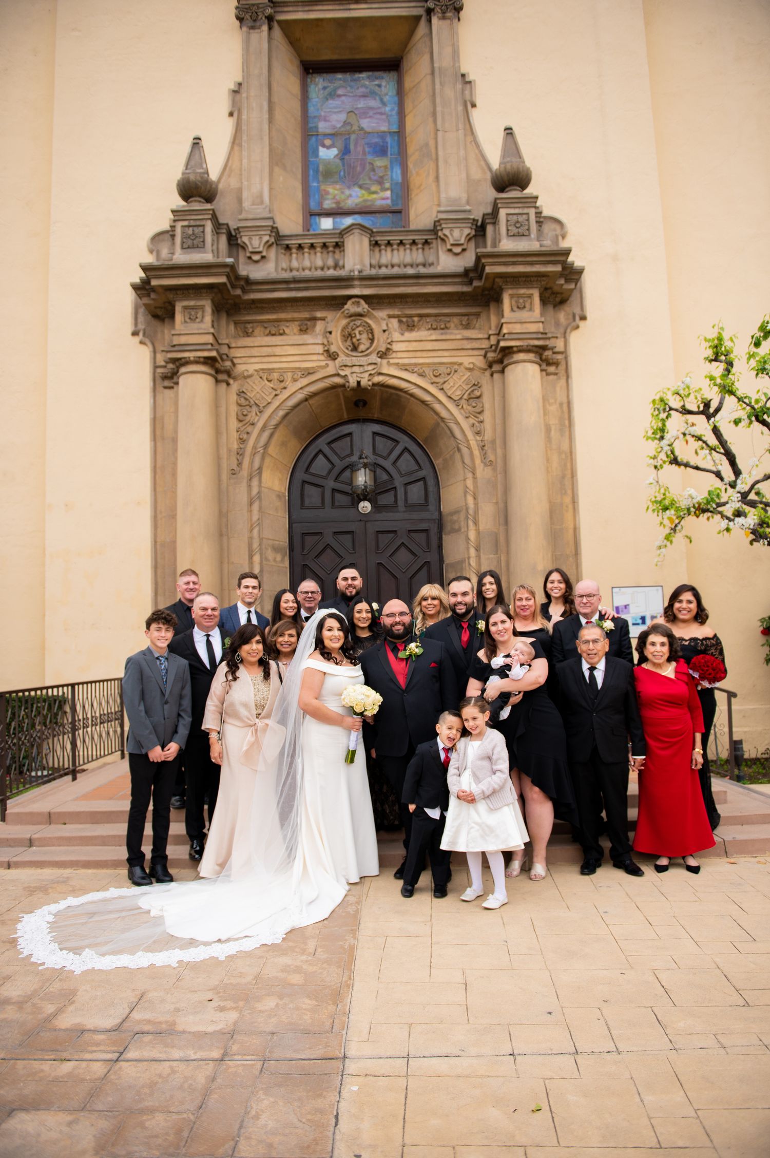 Wedding party poses on church steps in front of ornate stone archway with stained glass window above.