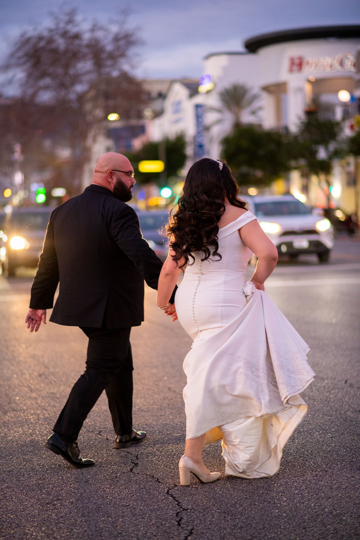 A couple in wedding attire walk through a city street at dusk with the bride's white dress flowing behind them.