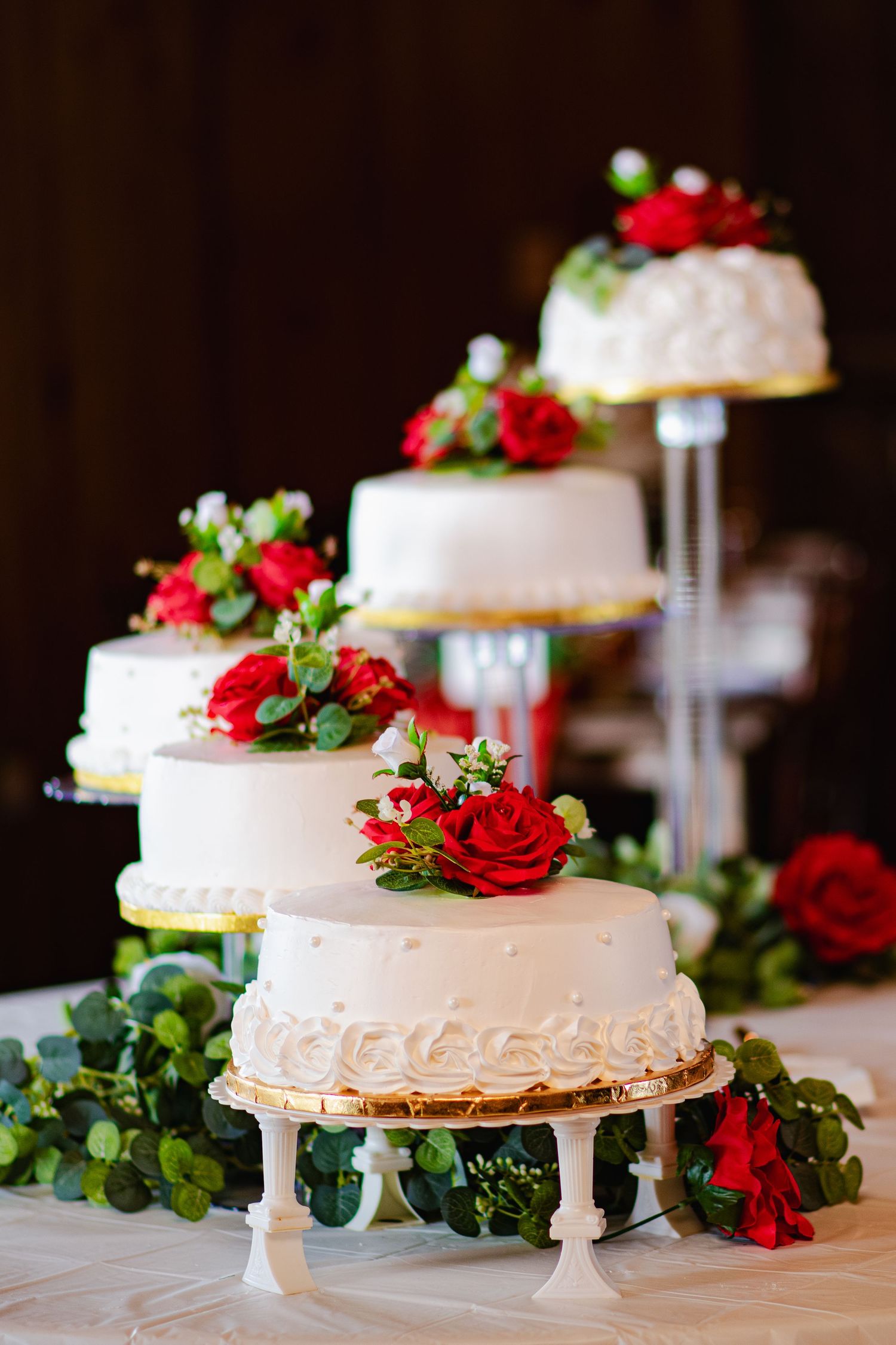 White wedding cake decorated with red roses and greenery displayed on elegant cake stands at reception.