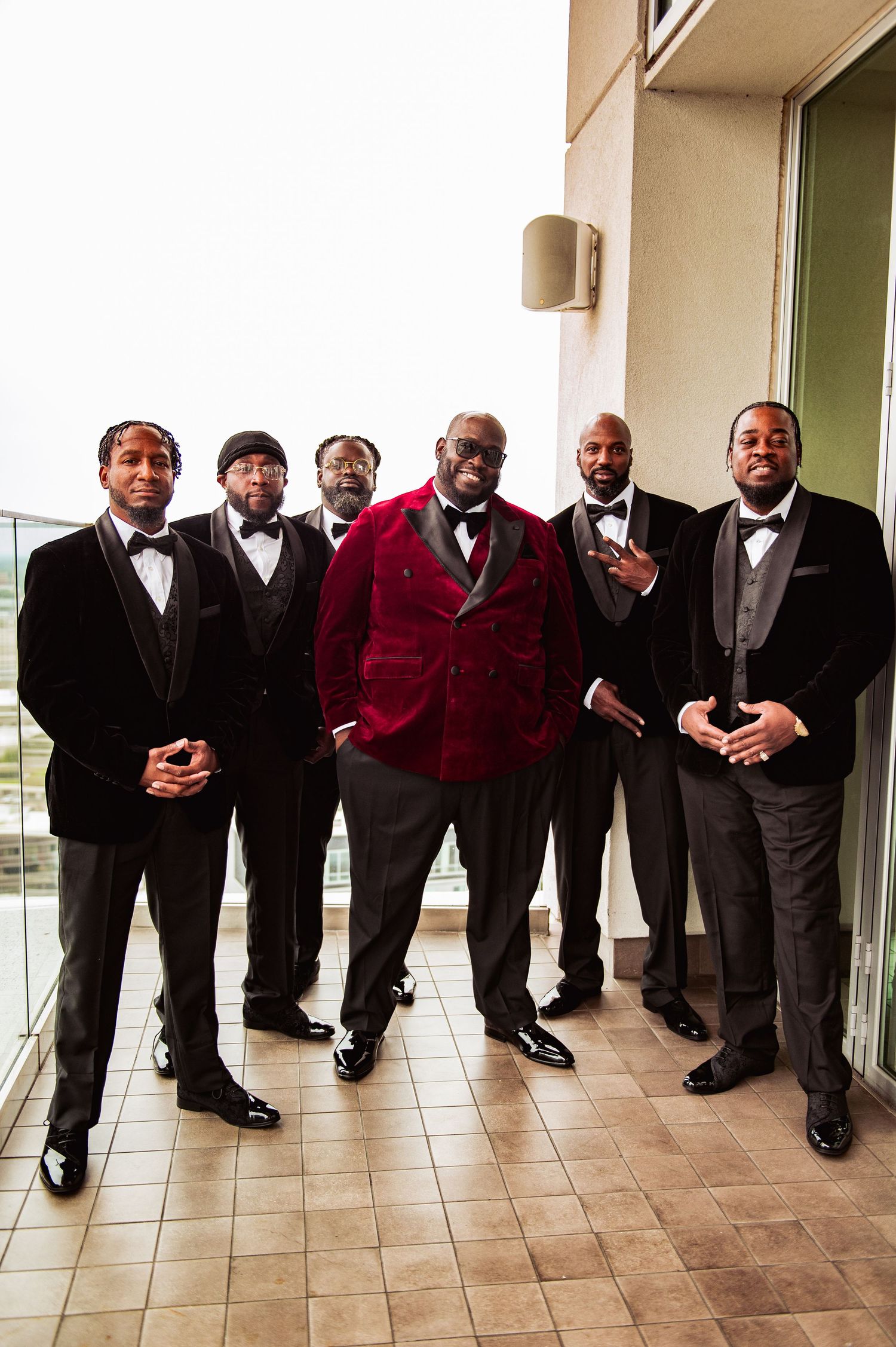 Groomsmen in formal black and burgundy tuxedos pose in a stylish lineup along a tiled hallway.