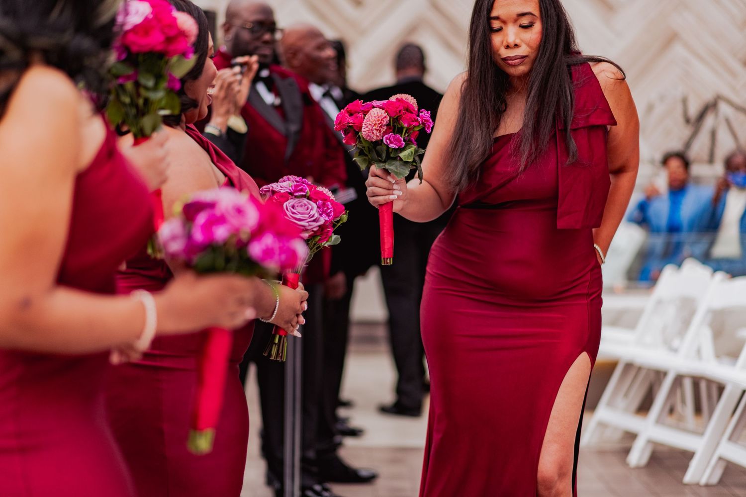 Bridesmaids in burgundy dresses hold vibrant pink and red bouquets during an elegant wedding ceremony.