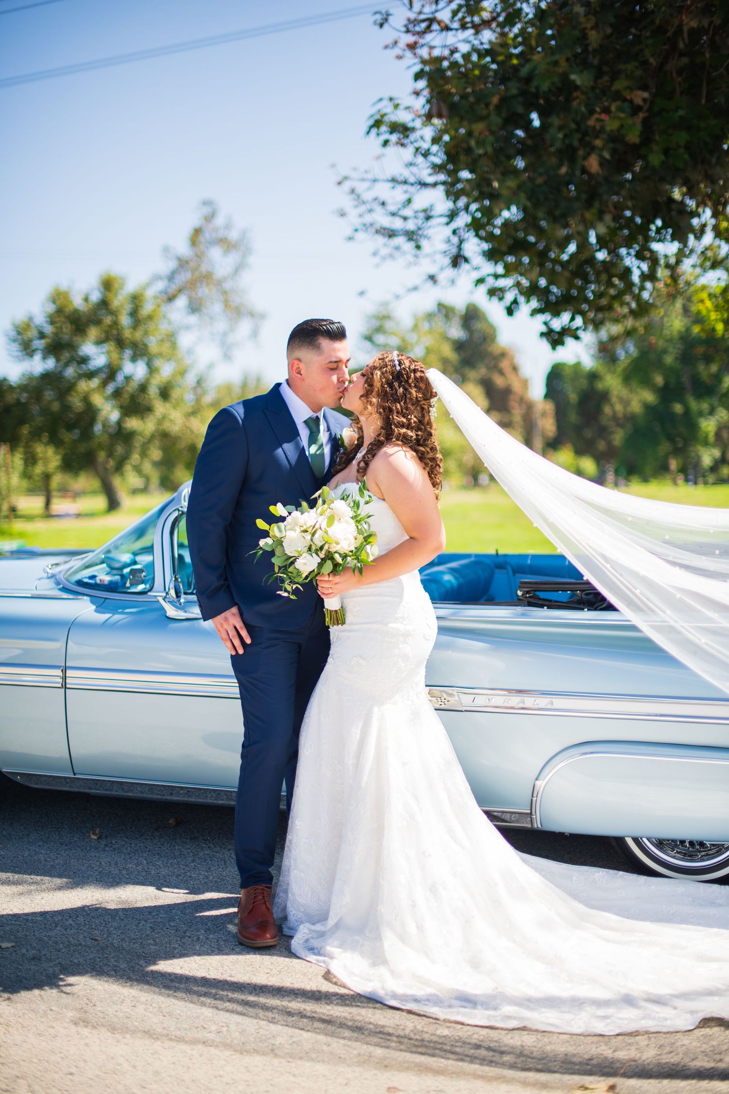 Newlyweds share intimate moment beside classic car with flowing white veil and bouquet.