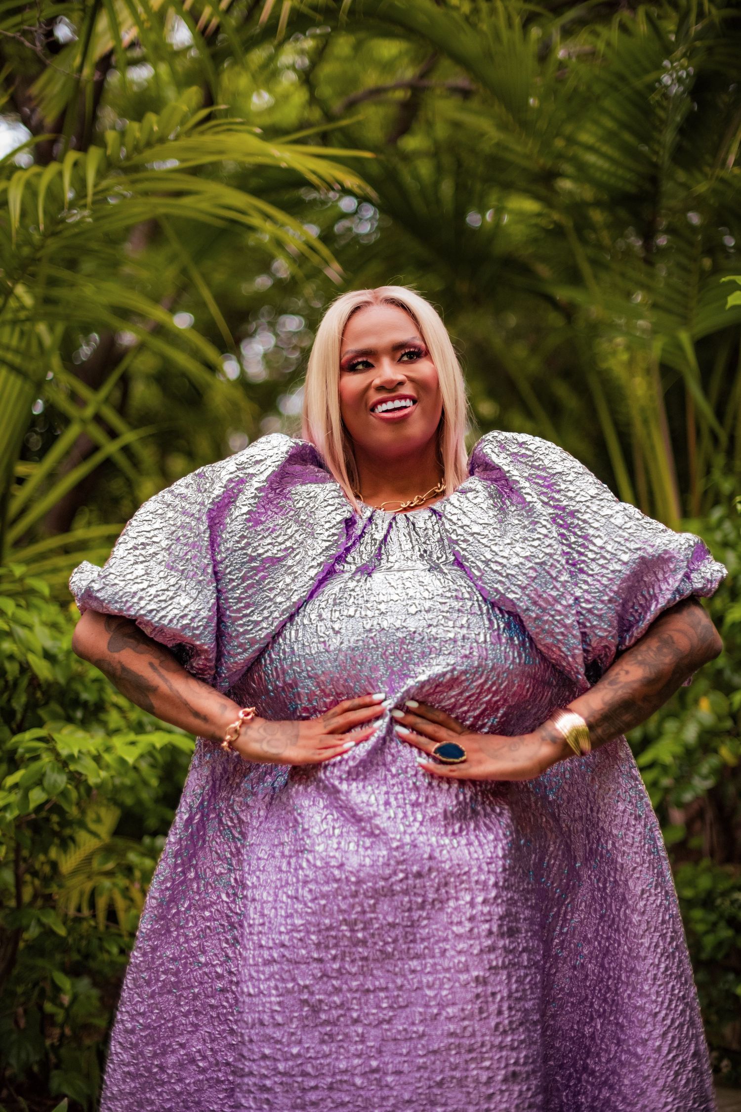 A person in a shimmering purple gown poses confidently against a backdrop of lush tropical foliage.