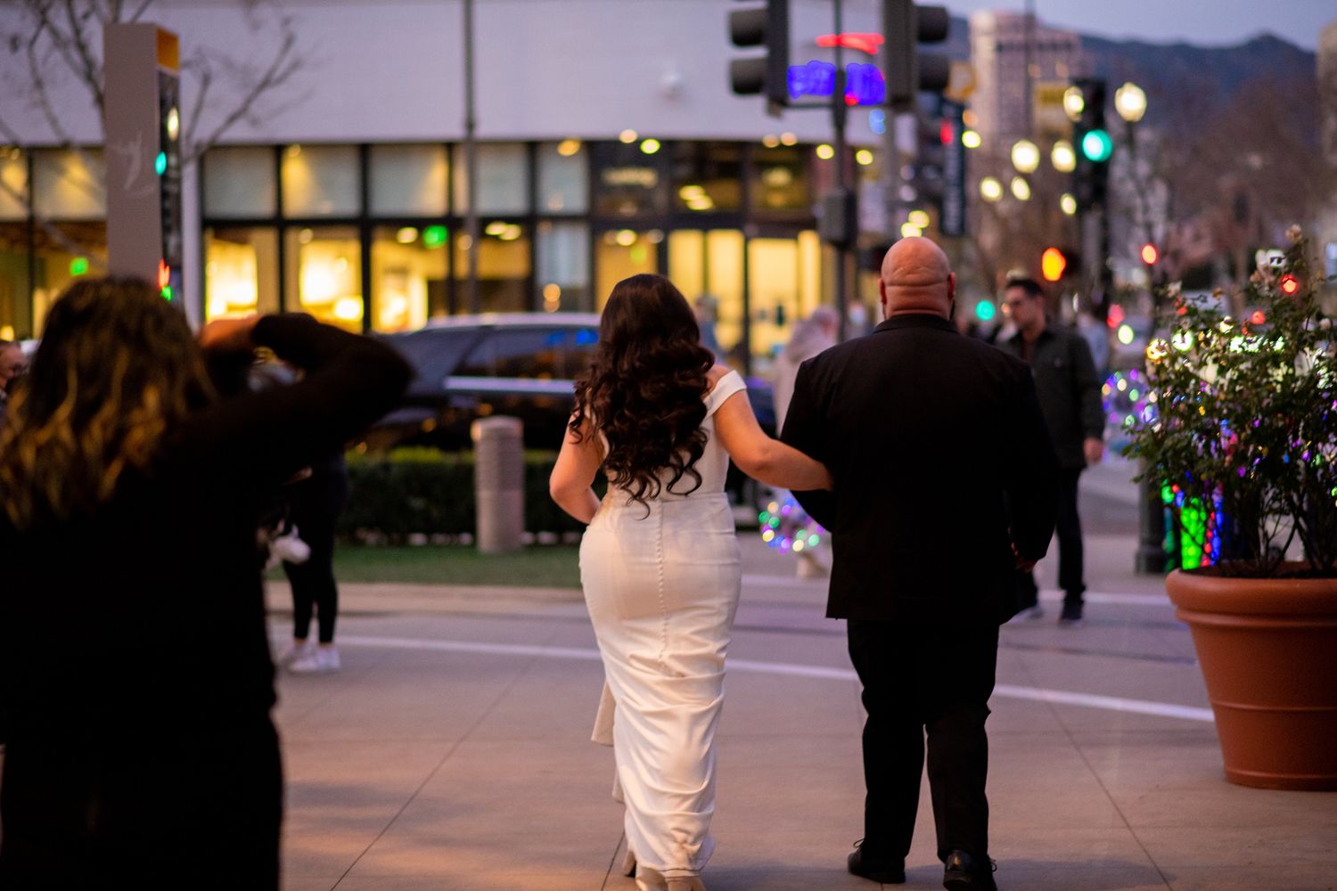People walk near illuminated storefronts in an outdoor shopping area at dusk with twinkling lights.