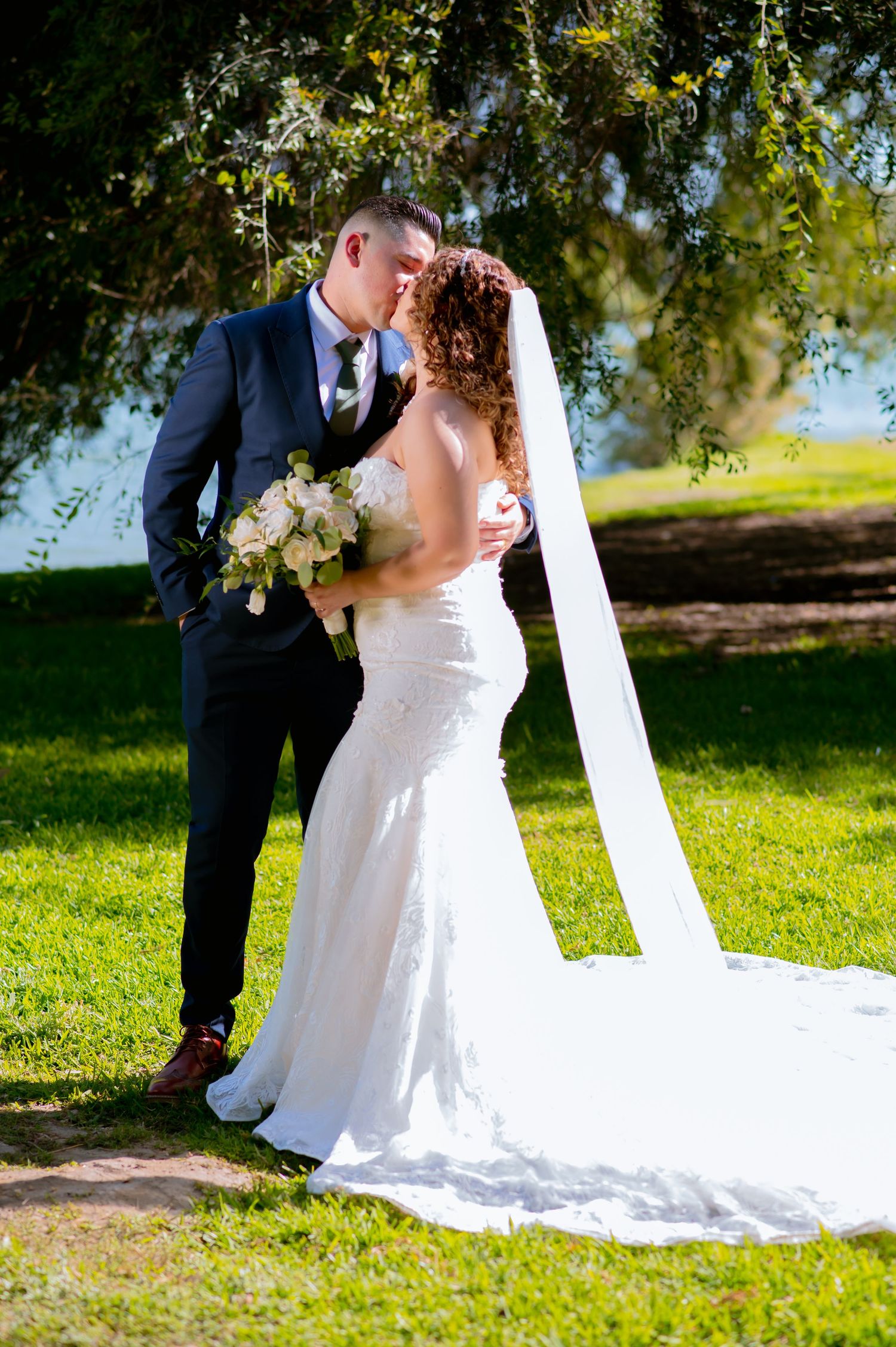 Wedding couple embracing on grassy lawn with flowing white veil and elegant attire.