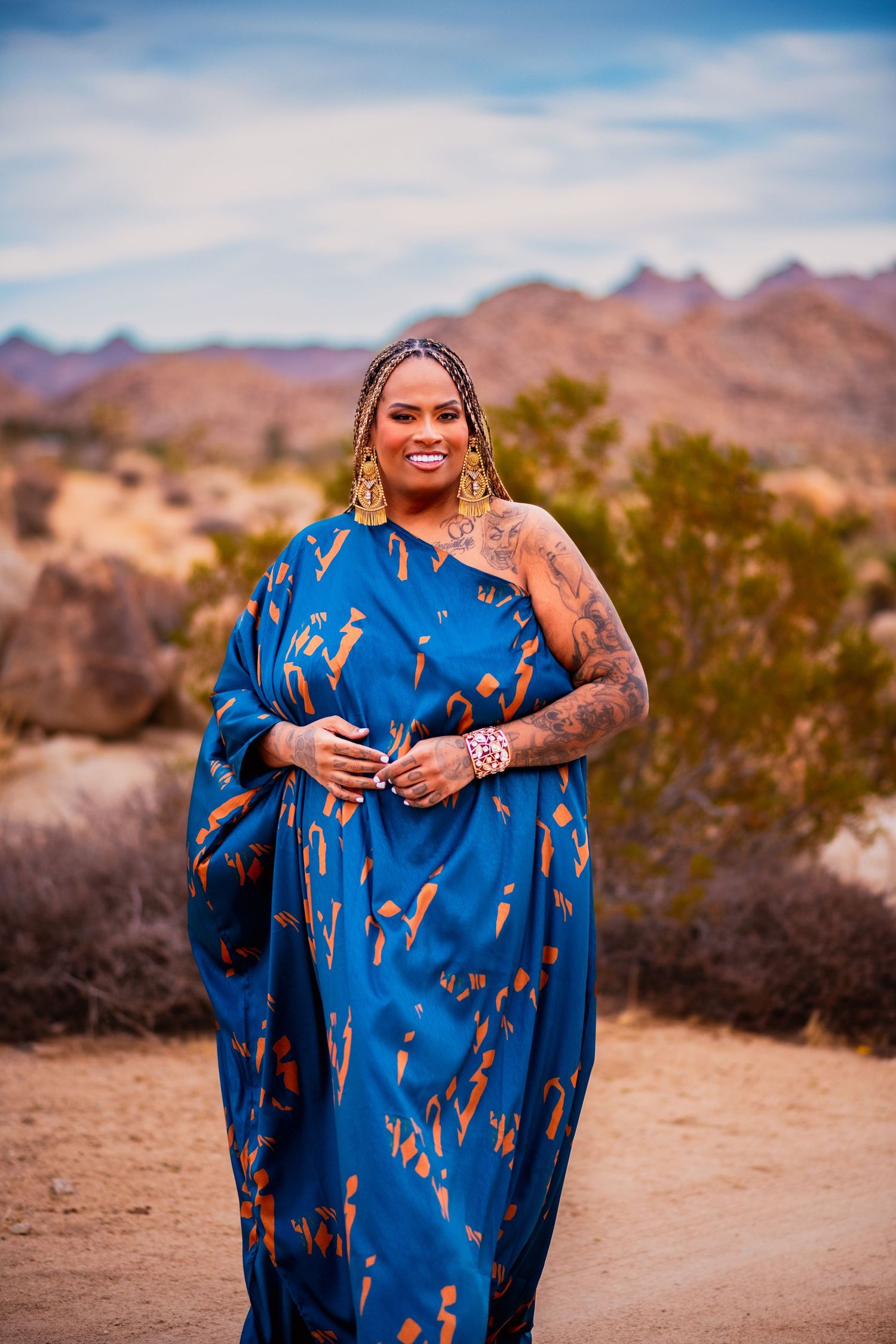 Fashion model poses in bright blue patterned dress against desert mountain scenery.