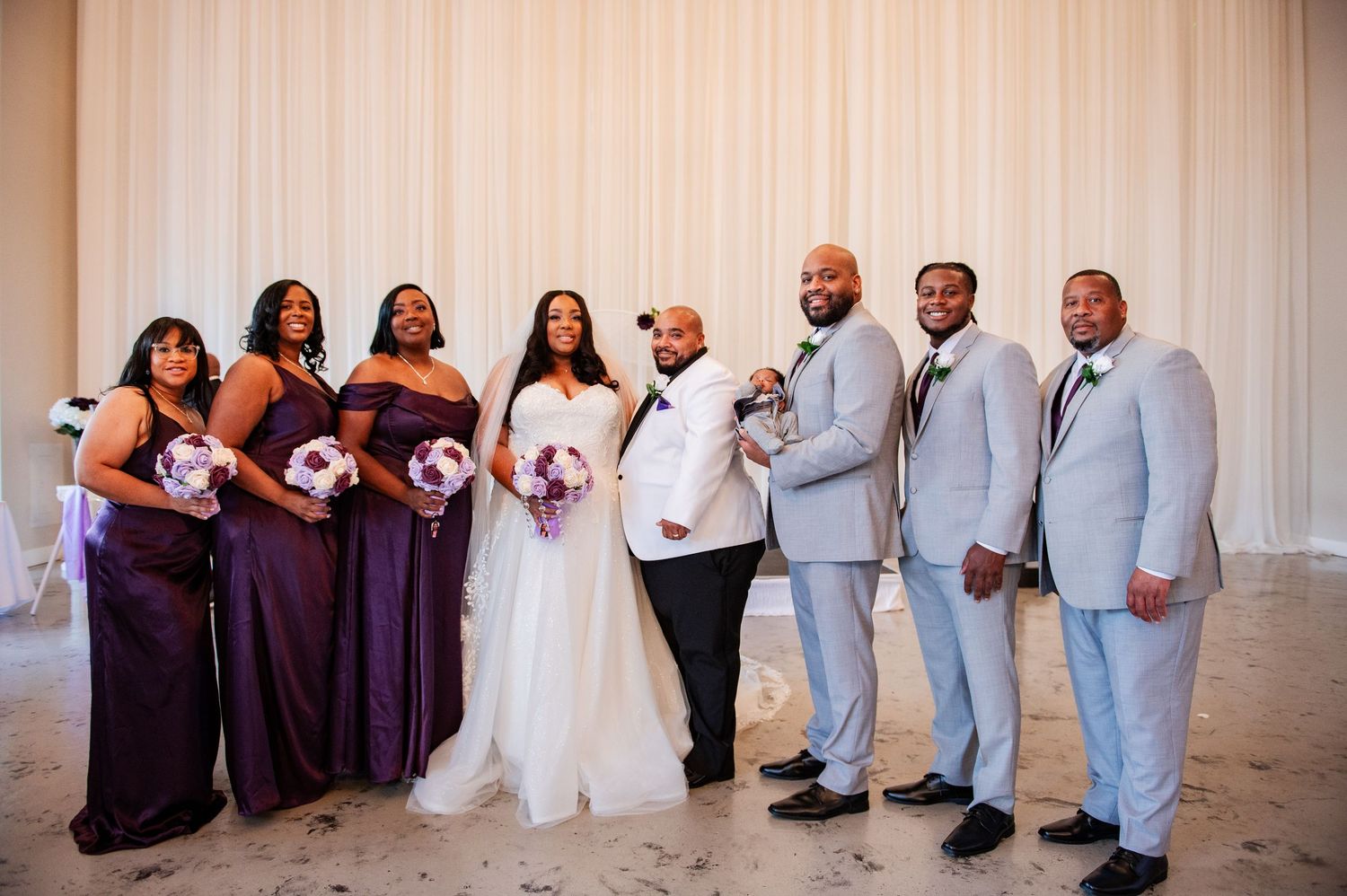 Wedding party lineup wearing purple dresses and gray suits in front of white backdrop.