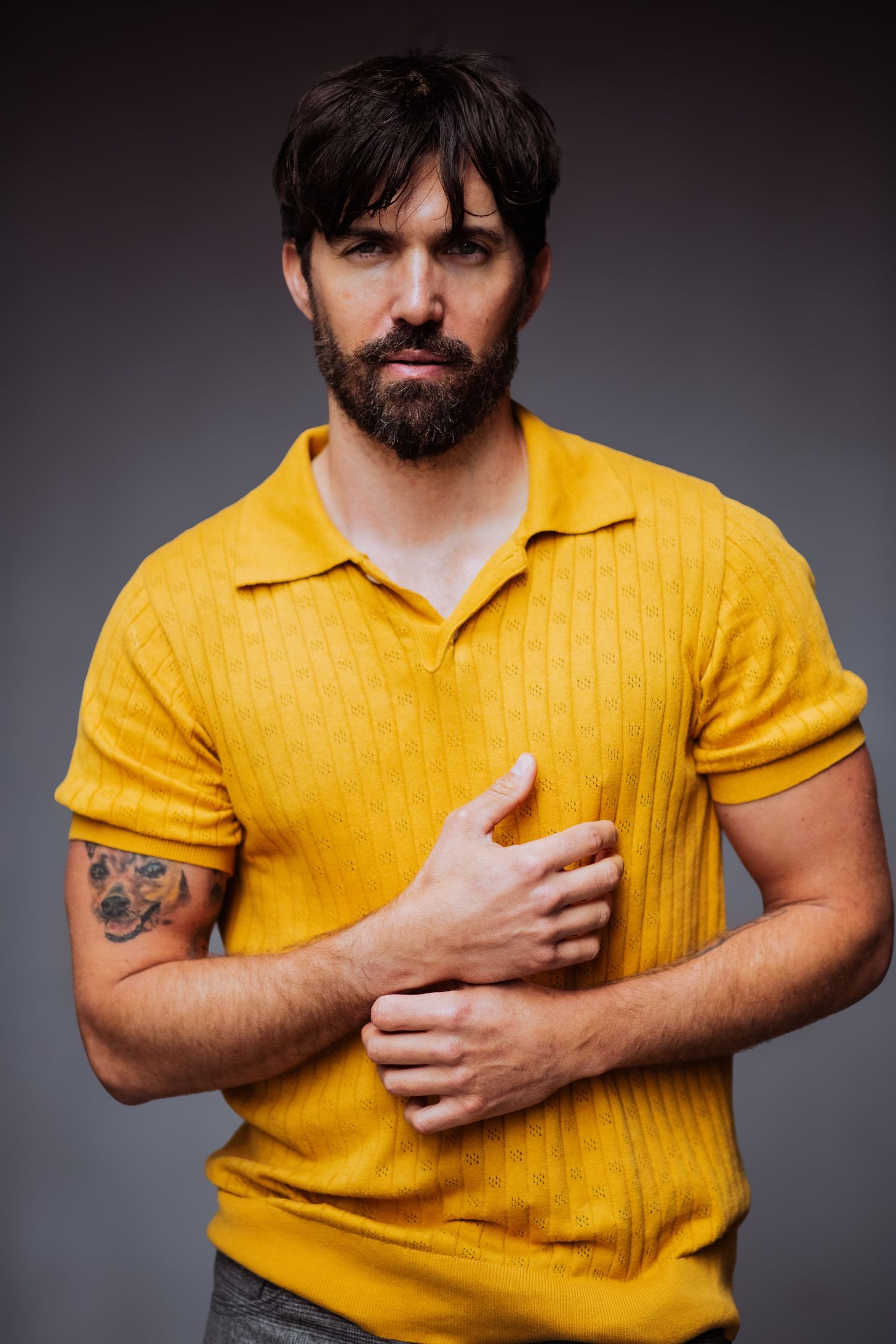 Individual in yellow collared shirt gestures with hand against chest in dramatic studio lighting.