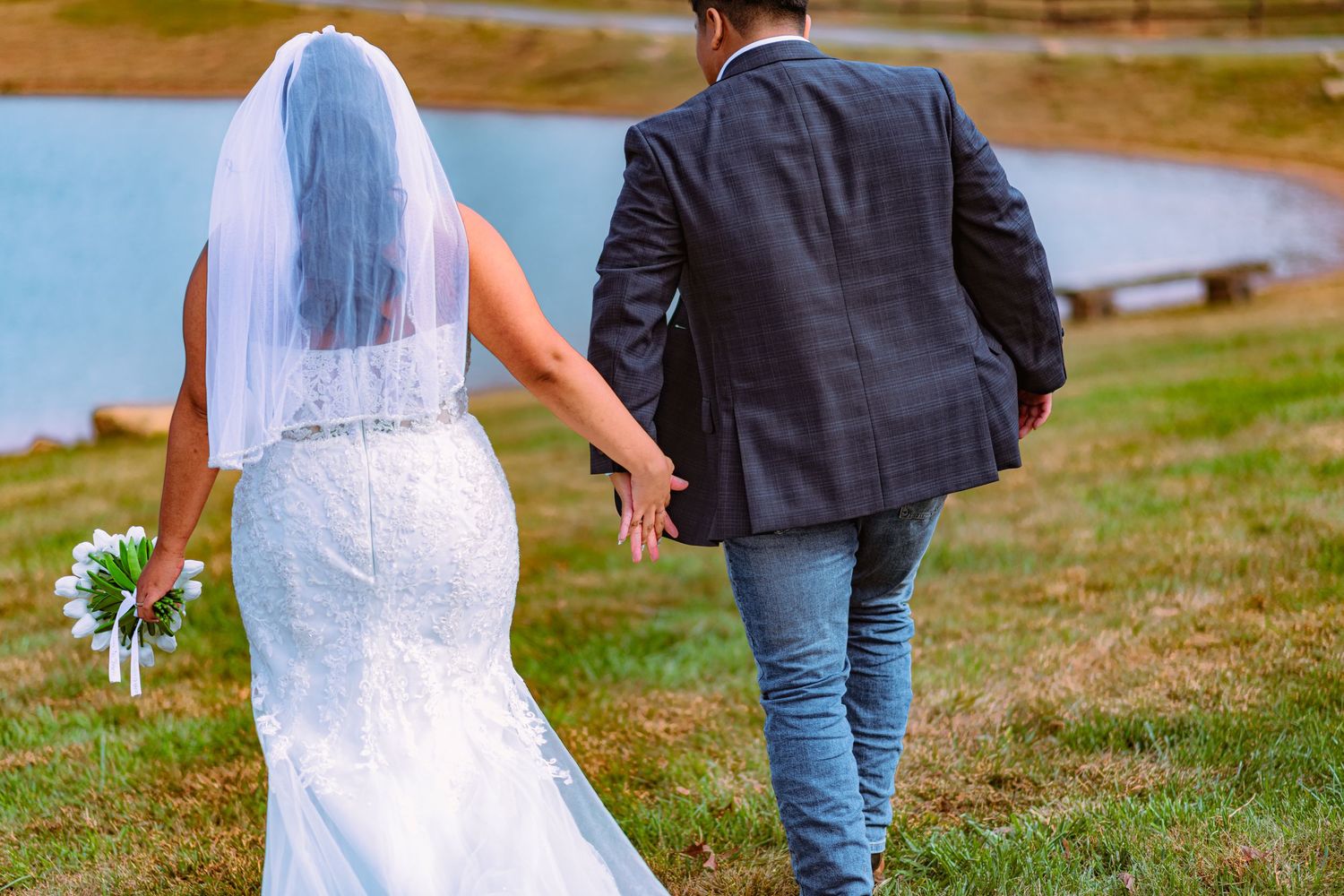 Couple walks hand in hand across grassy field during outdoor wedding celebration.