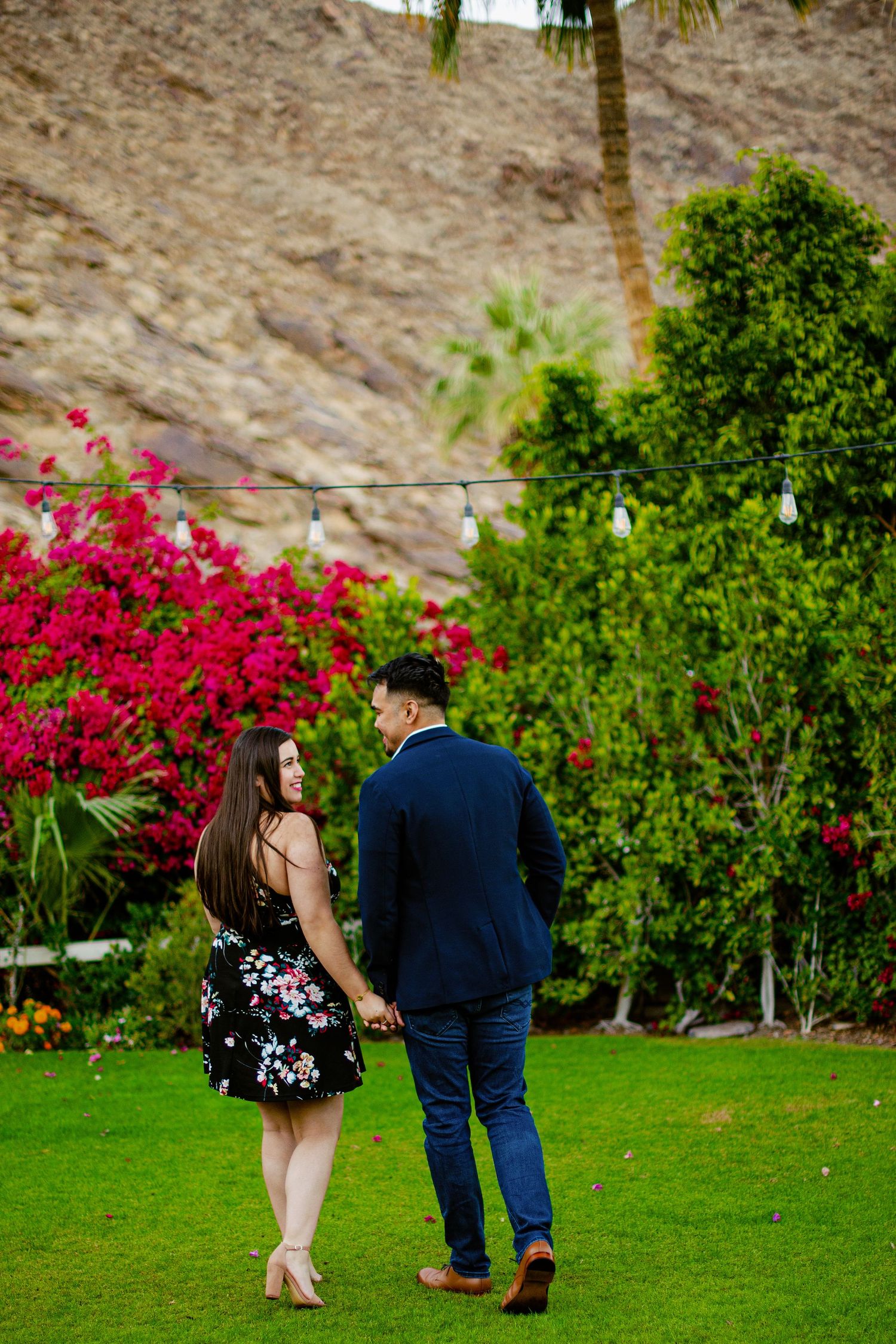 A couple shares a romantic moment on a green lawn with vibrant pink bougainvillea and lush foliage in the background.