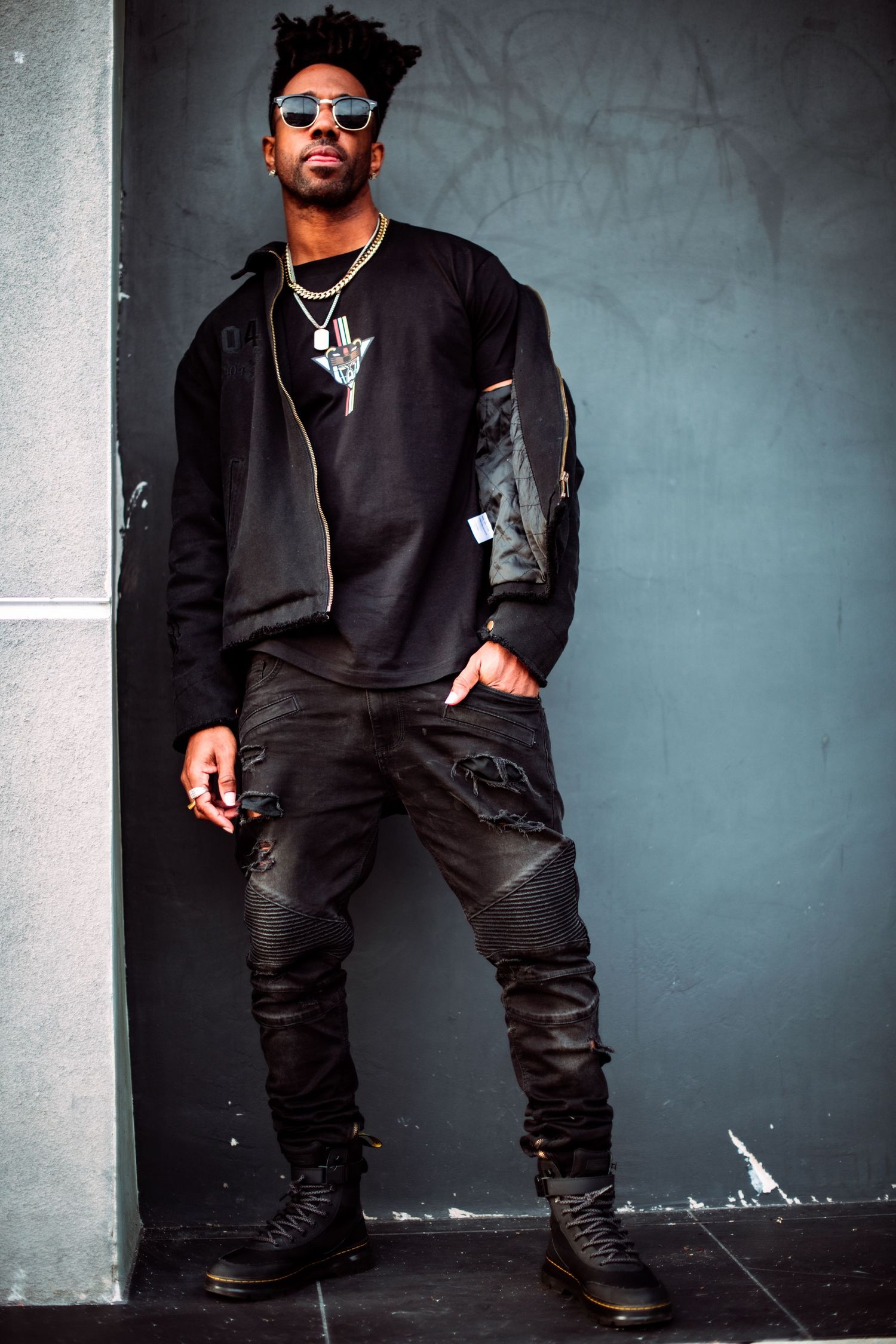 Cool urban fashion look with black bomber jacket, distressed jeans, boots, sunglasses and silver pendant against gray wall.
