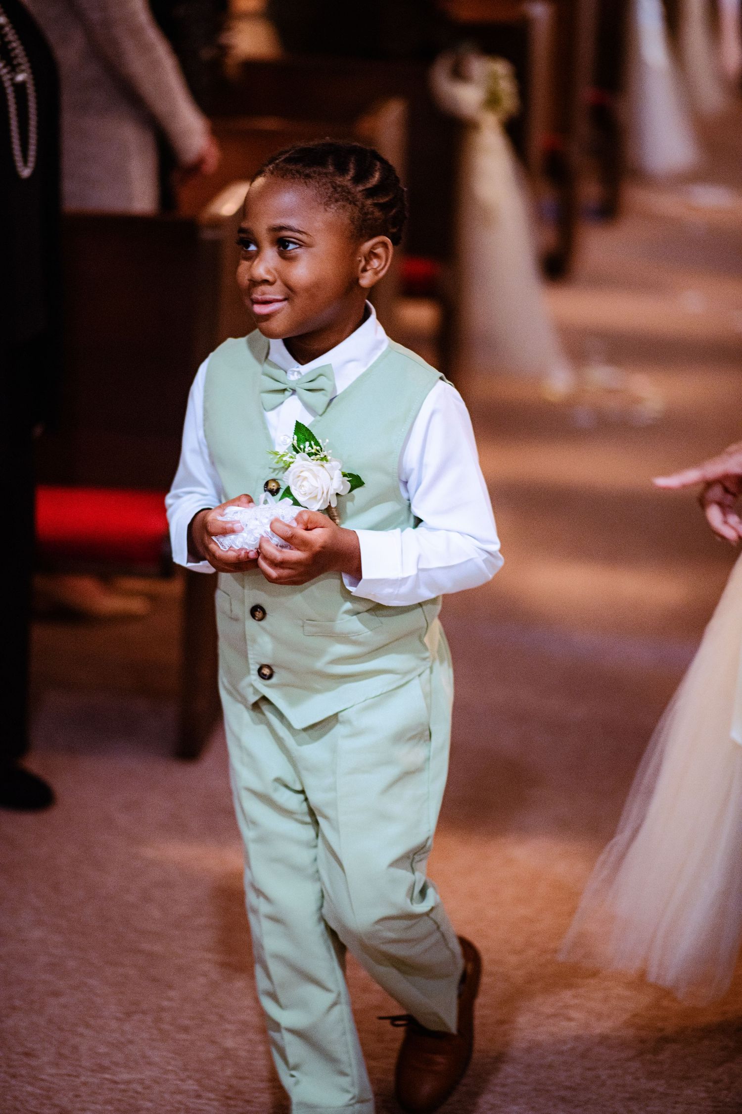 A young attendant in a mint green suit carries flowers during a wedding ceremony.